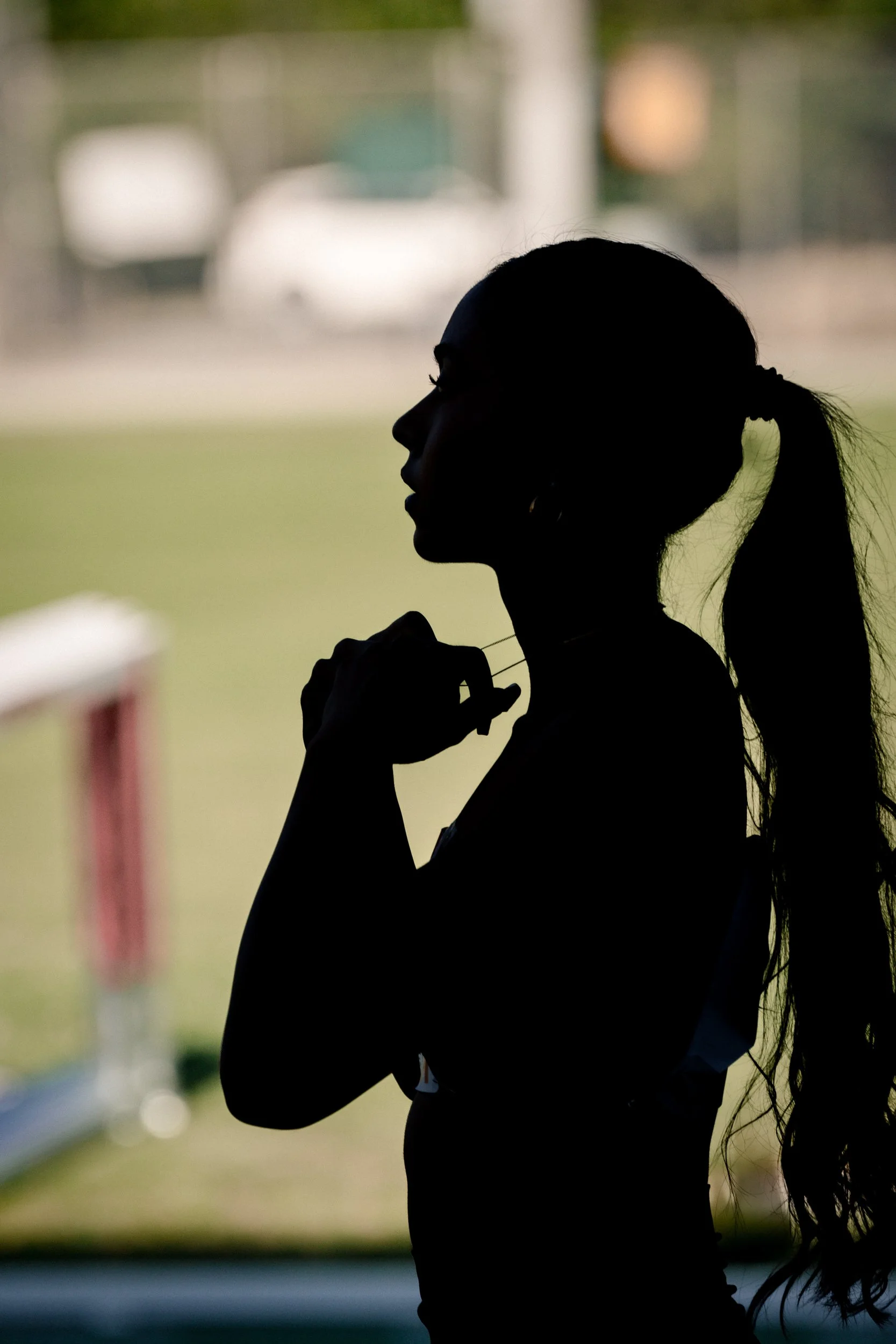 Silhouette of a woman with a ponytail, holding earbuds near her neck, in front of a blurred outdoor background.