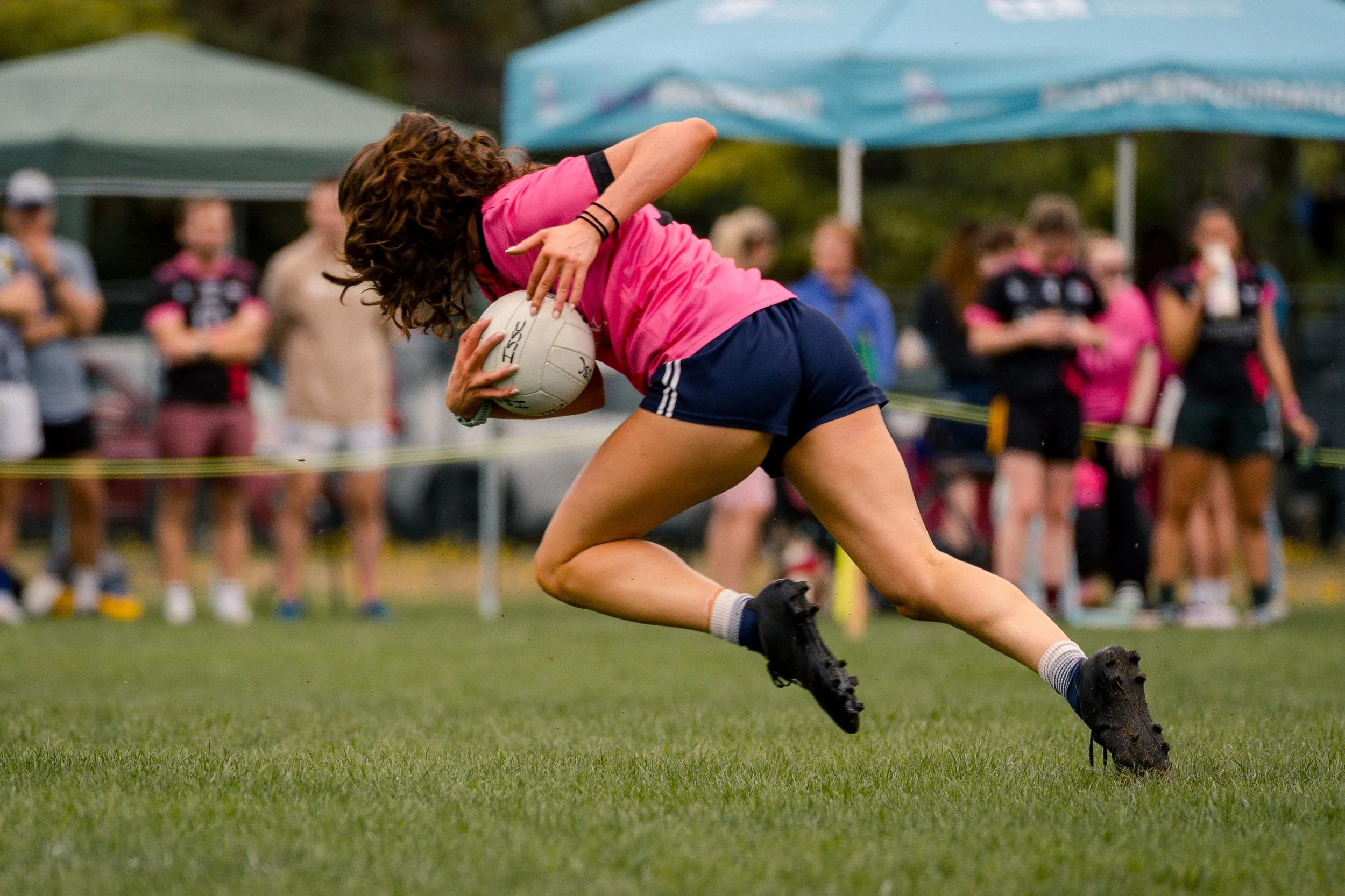 A woman playing rugby, holding a rugby ball while running on a grassy field. She is wearing a pink shirt, navy shorts, and black cleats, with a group of people watching in the background.