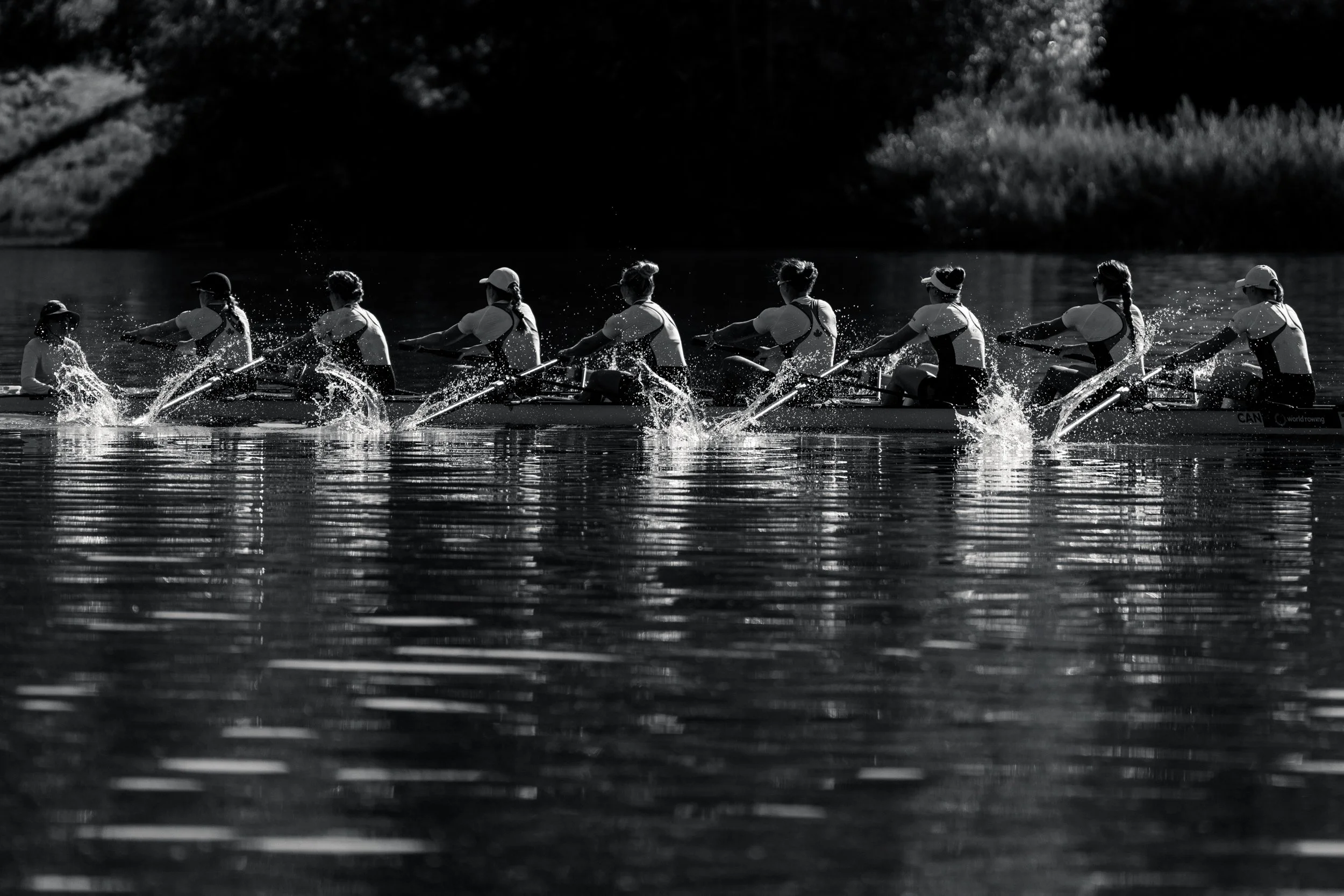 Eight rowers in a boat, paddling on a river during daytime, with splashes of water around their oars, in black and white.