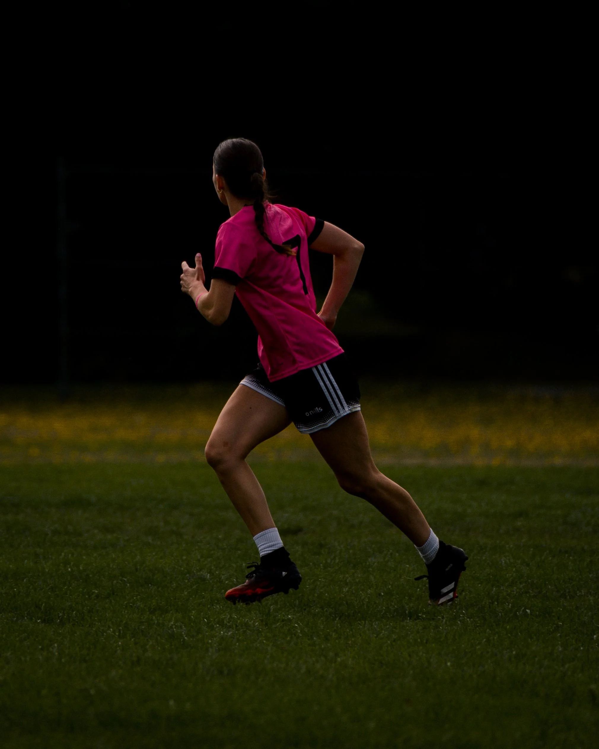 A female soccer player wearing a pink jersey, black shorts, and black cleats running on a grass field during night or evening.