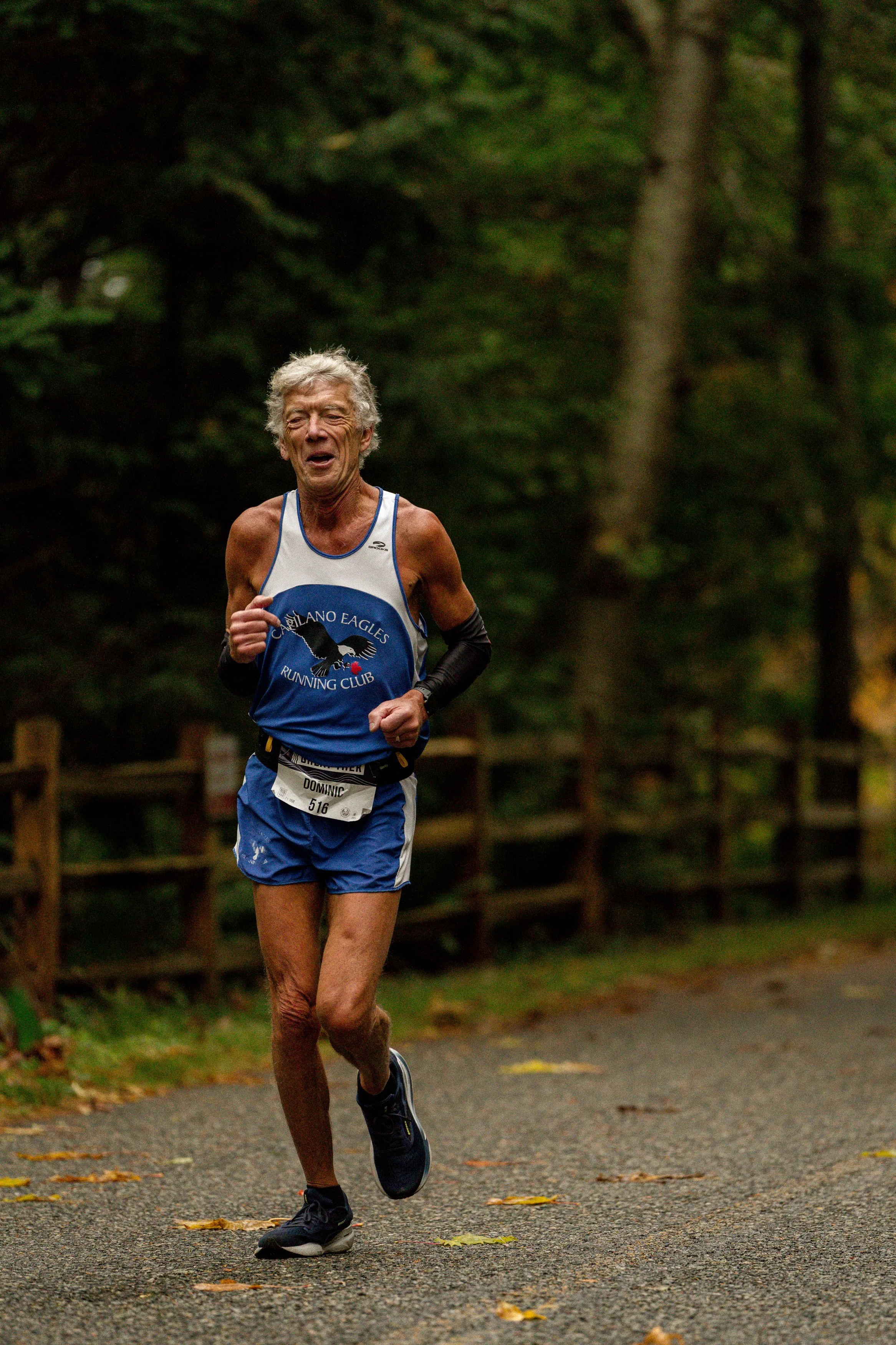 An elderly male runner with gray hair, wearing a blue and white tank top and shorts, is running on a forest trail during a race.