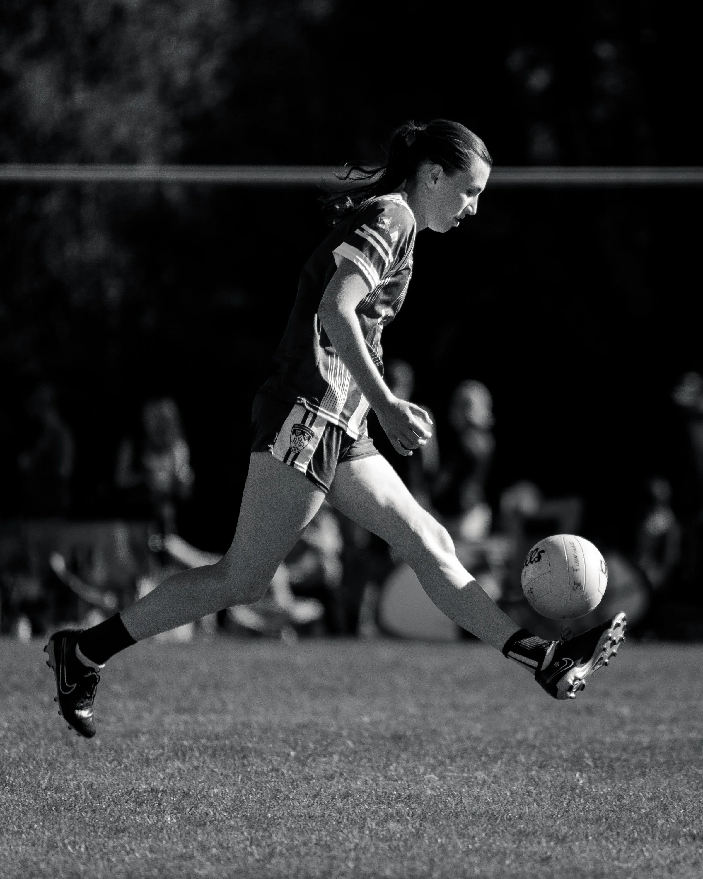 A female soccer player in a uniform kicks a soccer ball on the field during a game, with spectators watching in the background.