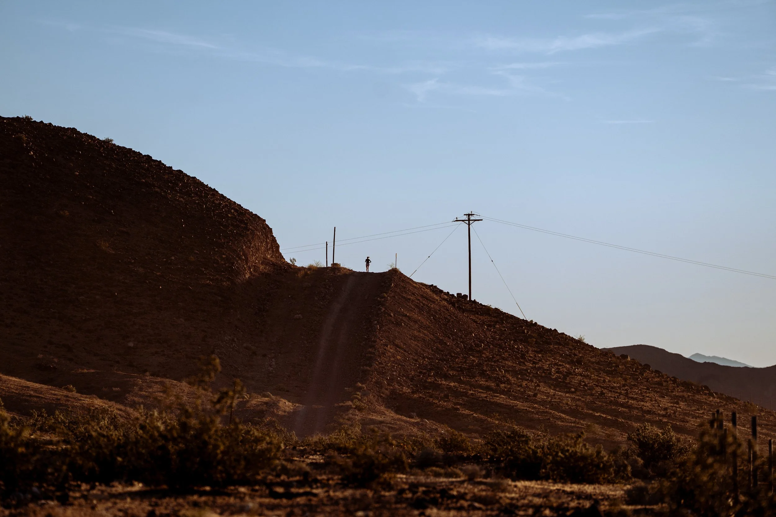 Hilly landscape at sunset with a person standing near power lines on a dirt path.