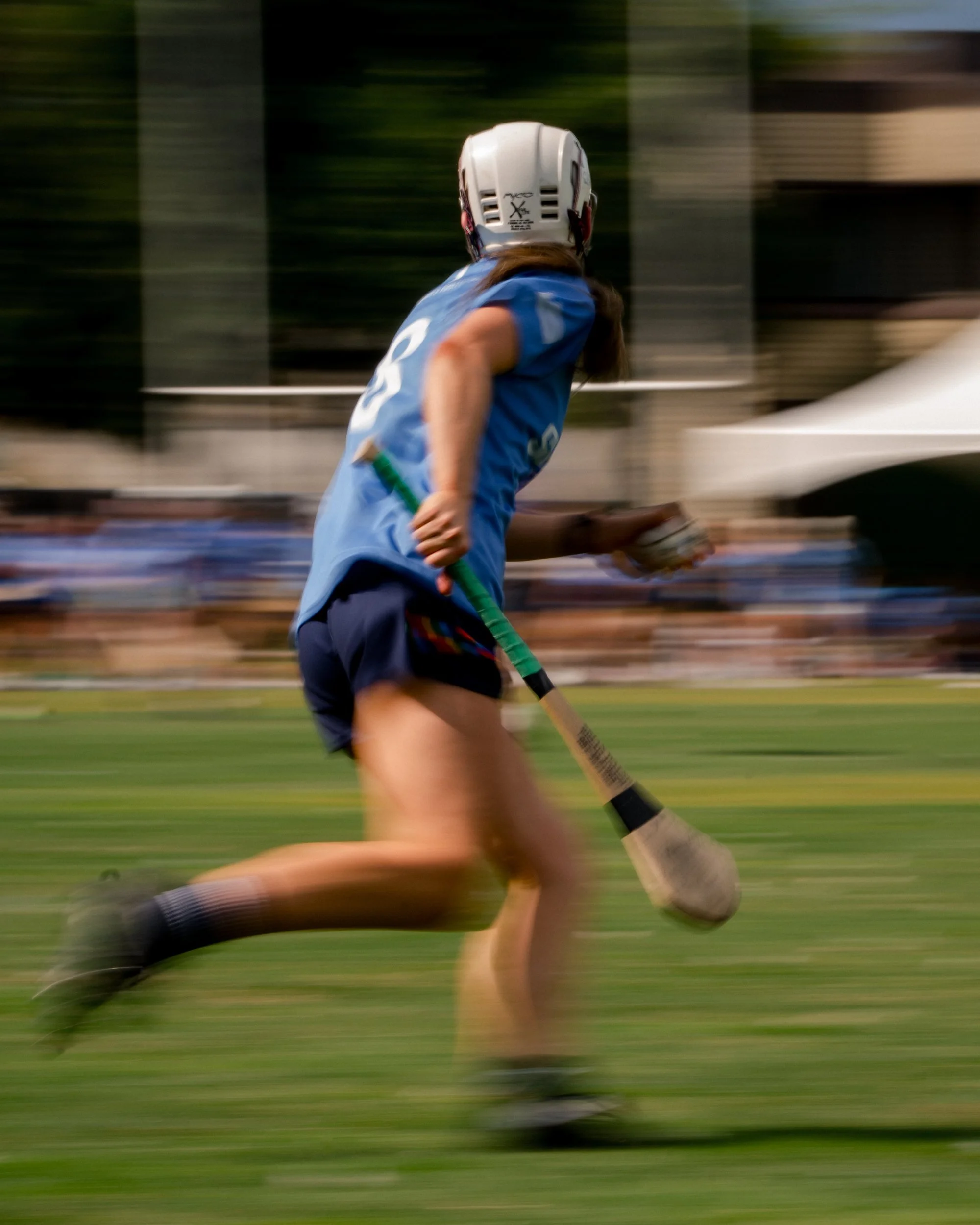 A female athlete in a blue jersey and shorts playing field hockey during a game, wearing a white helmet, running on the grass with a field hockey stick in her right hand.