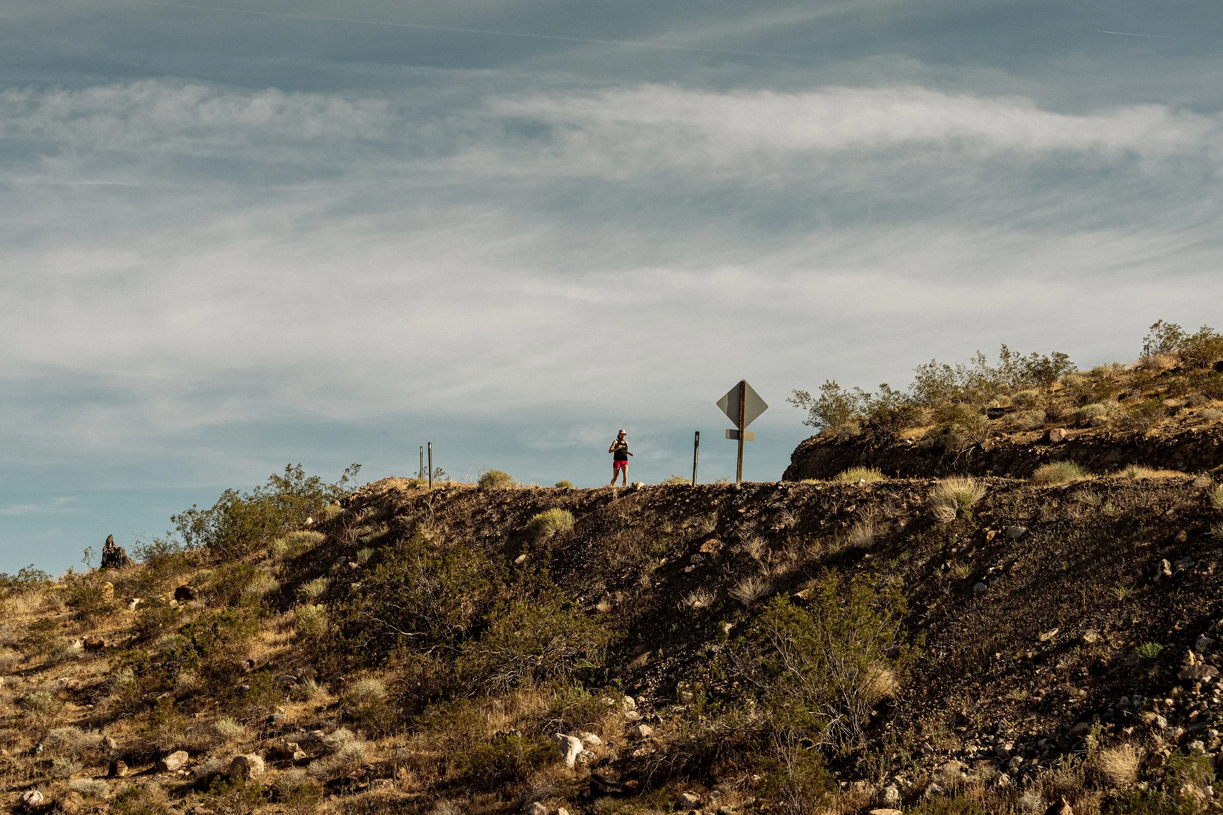 A person standing on a dry, rocky hillside, holding a camera or phone, with a cloudy sky above and sparse bushes and trees on the slope.