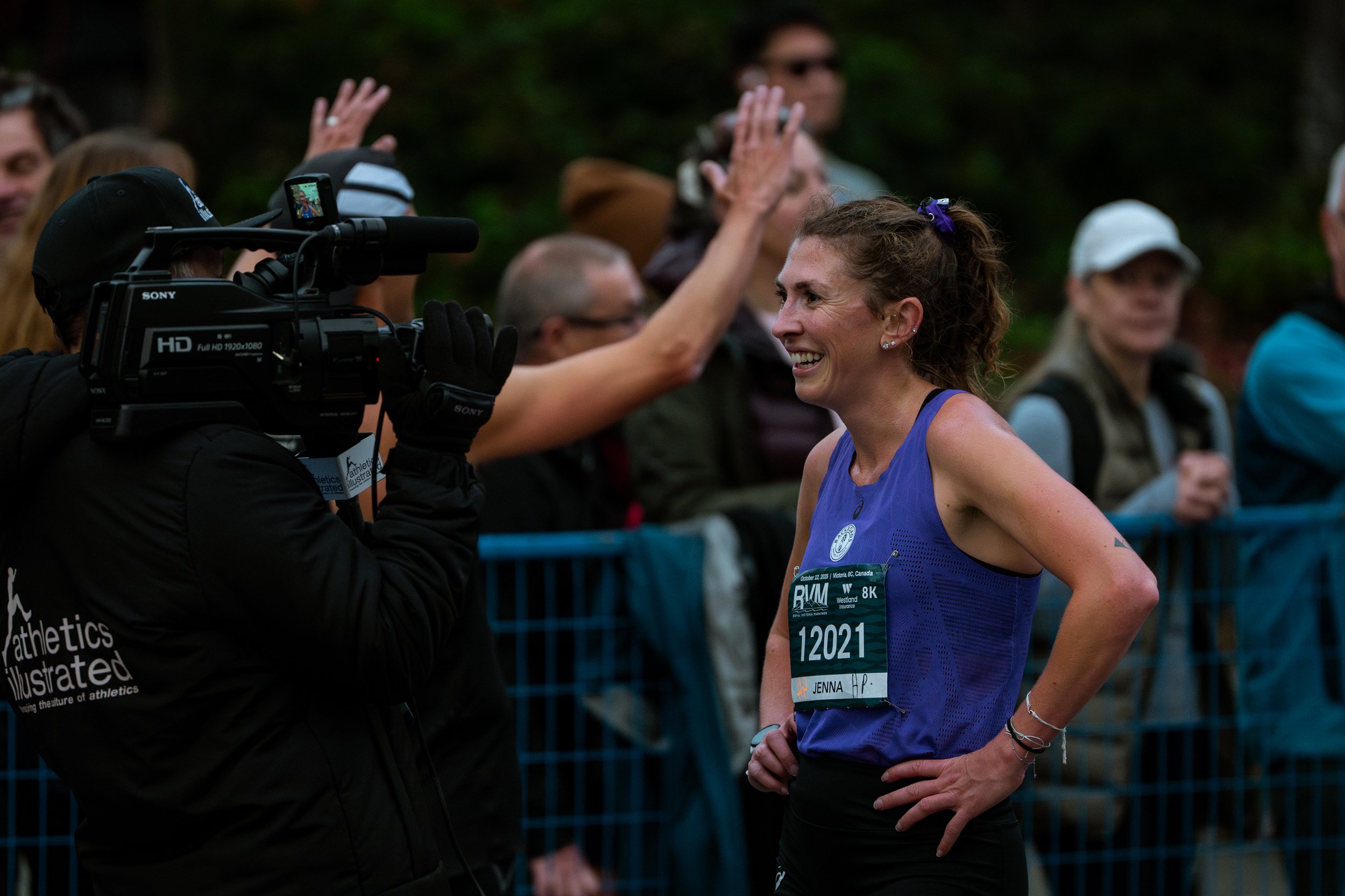 A female marathon runner being interviewed by a cameraman at a race event, with onlookers in the background.
