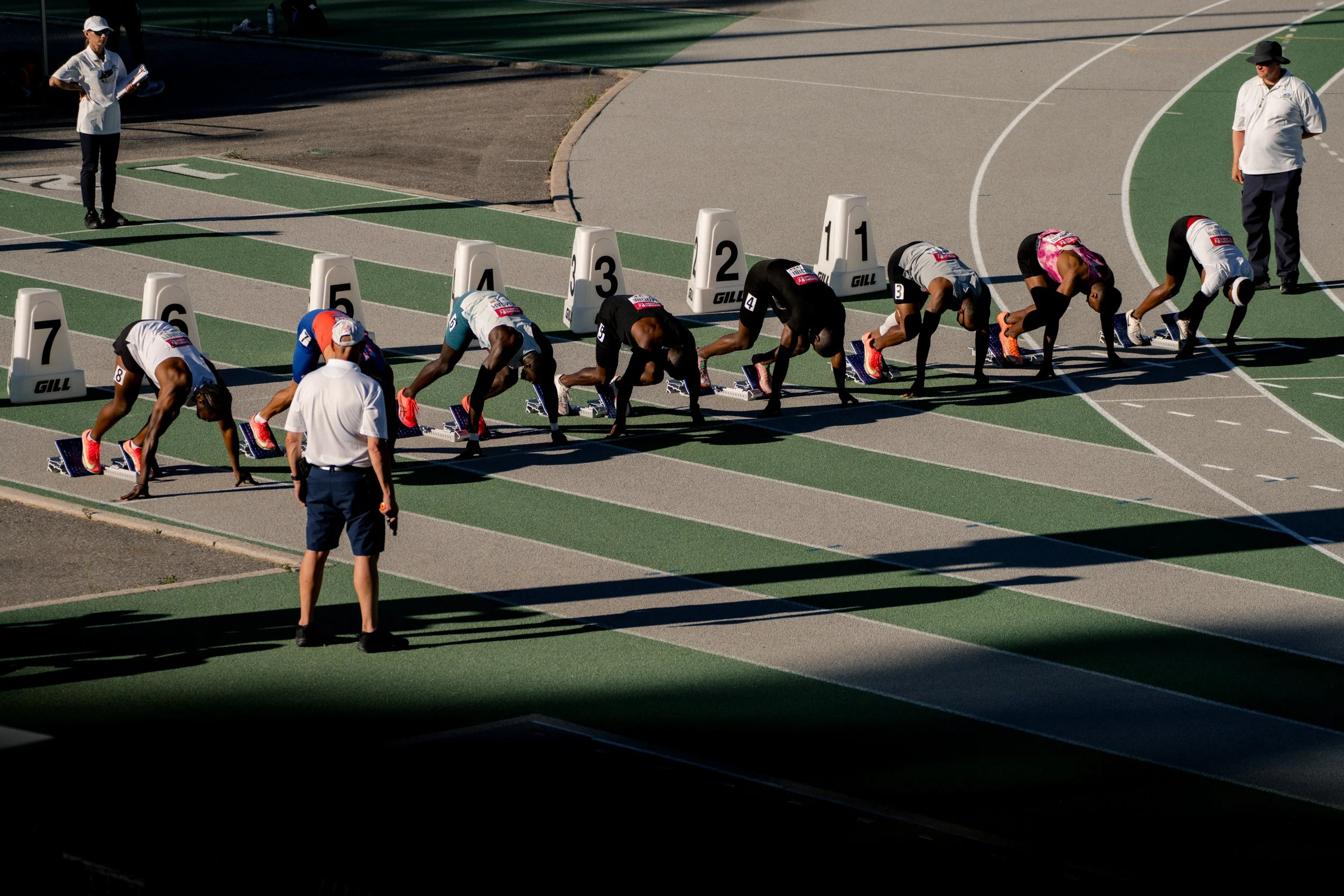 Track athletes starting a race with officials nearby on a green running track.