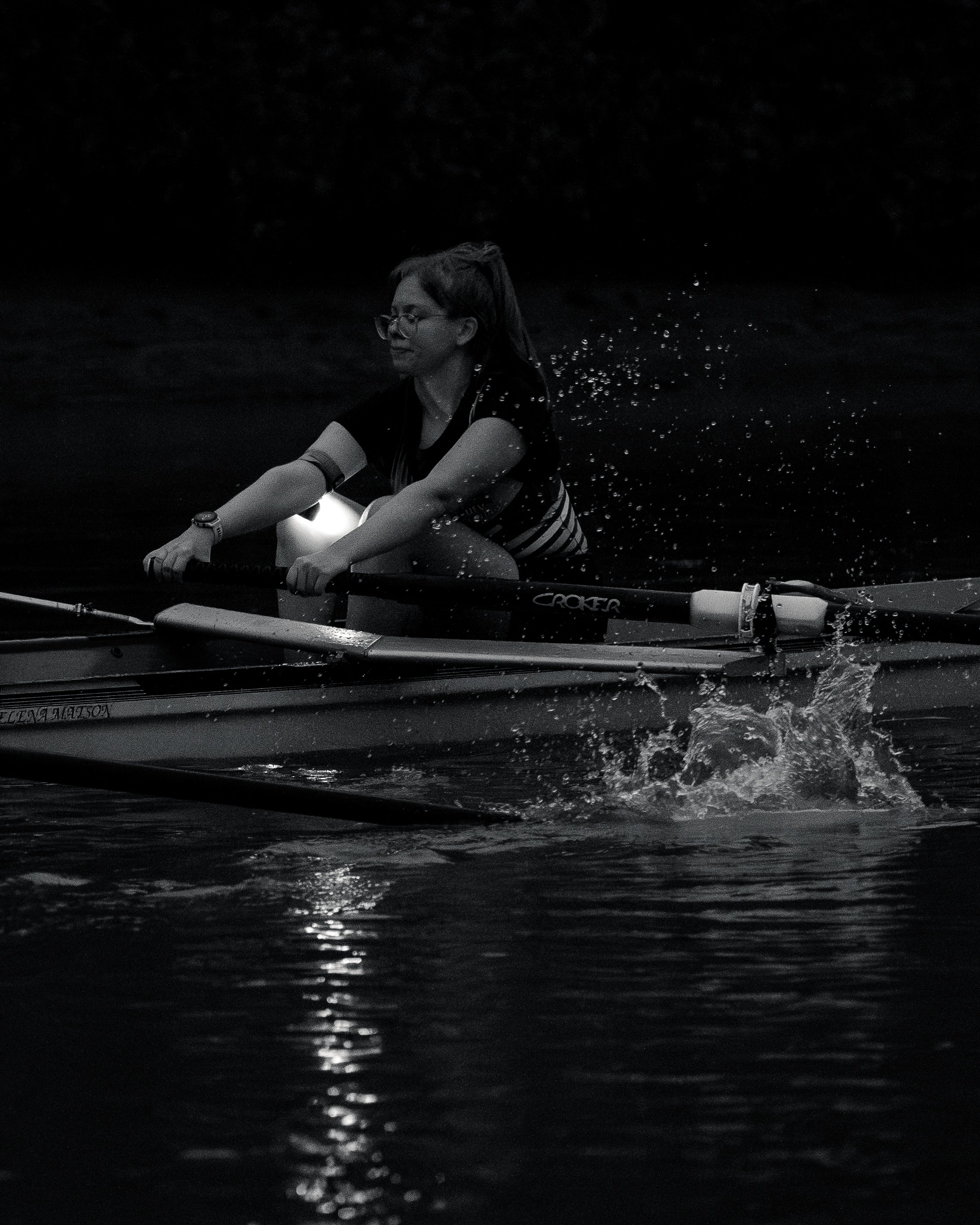 A woman with glasses paddling a canoe or kayak on dark water at night, with water splashing around her.