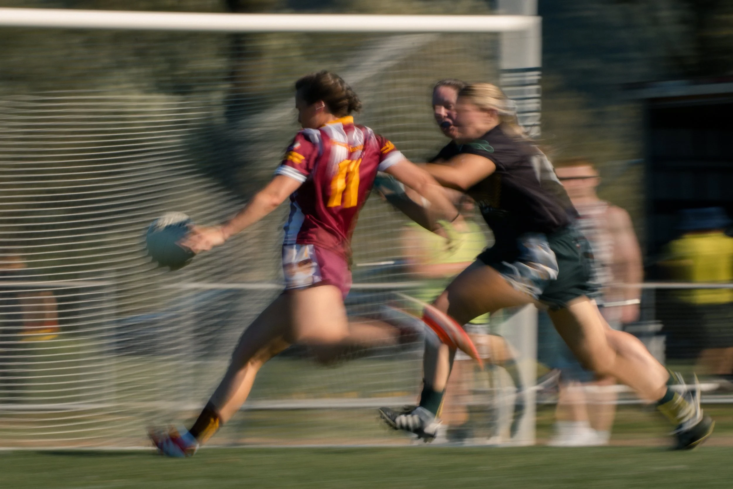 Rugby game with players running, one in maroon uniform holding the ball, pursued by others in black uniforms, with a netted background and spectators.