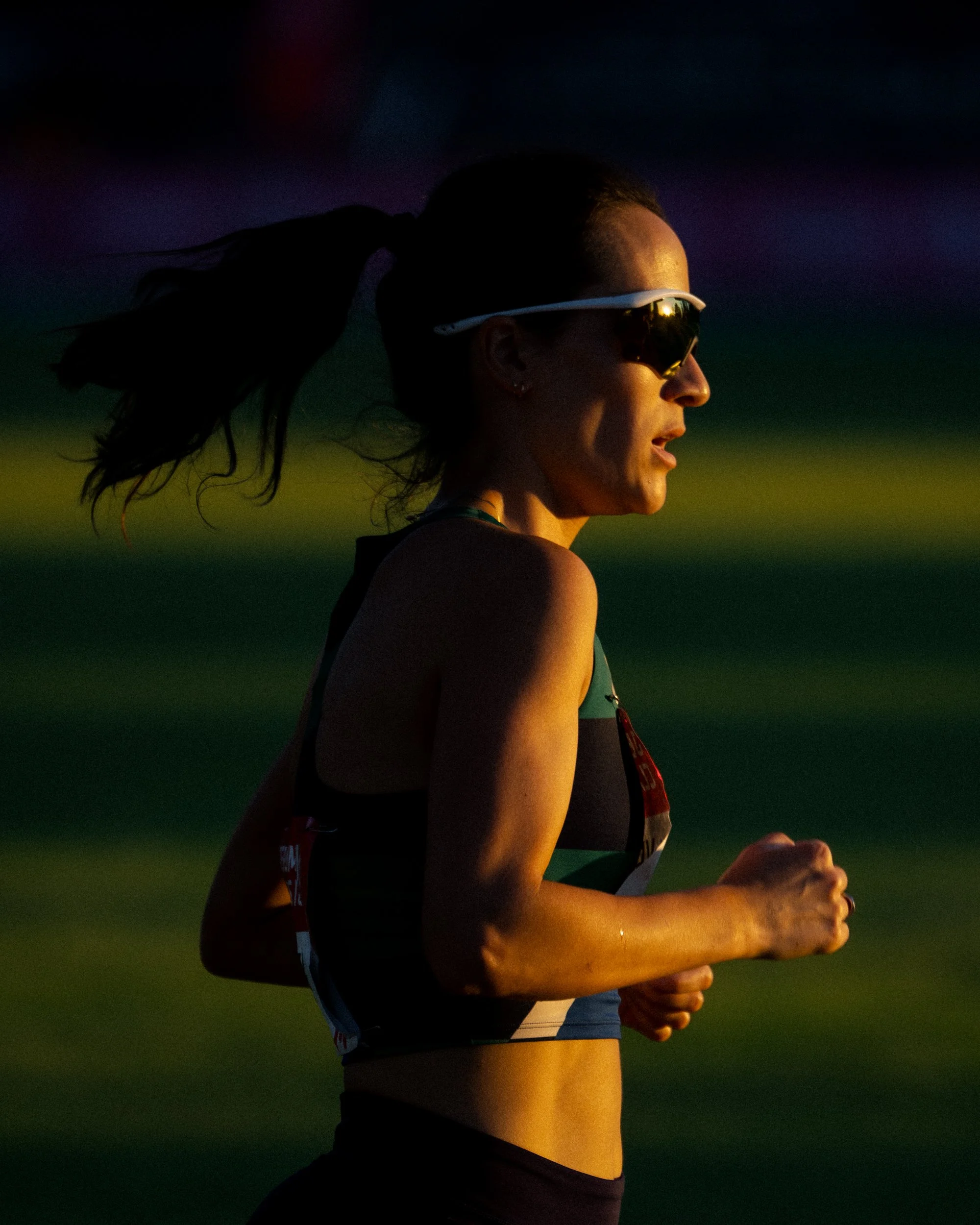 A female runner with dark hair tied back, wearing sunglasses and athletic gear, running outdoors during sunset or sunrise.