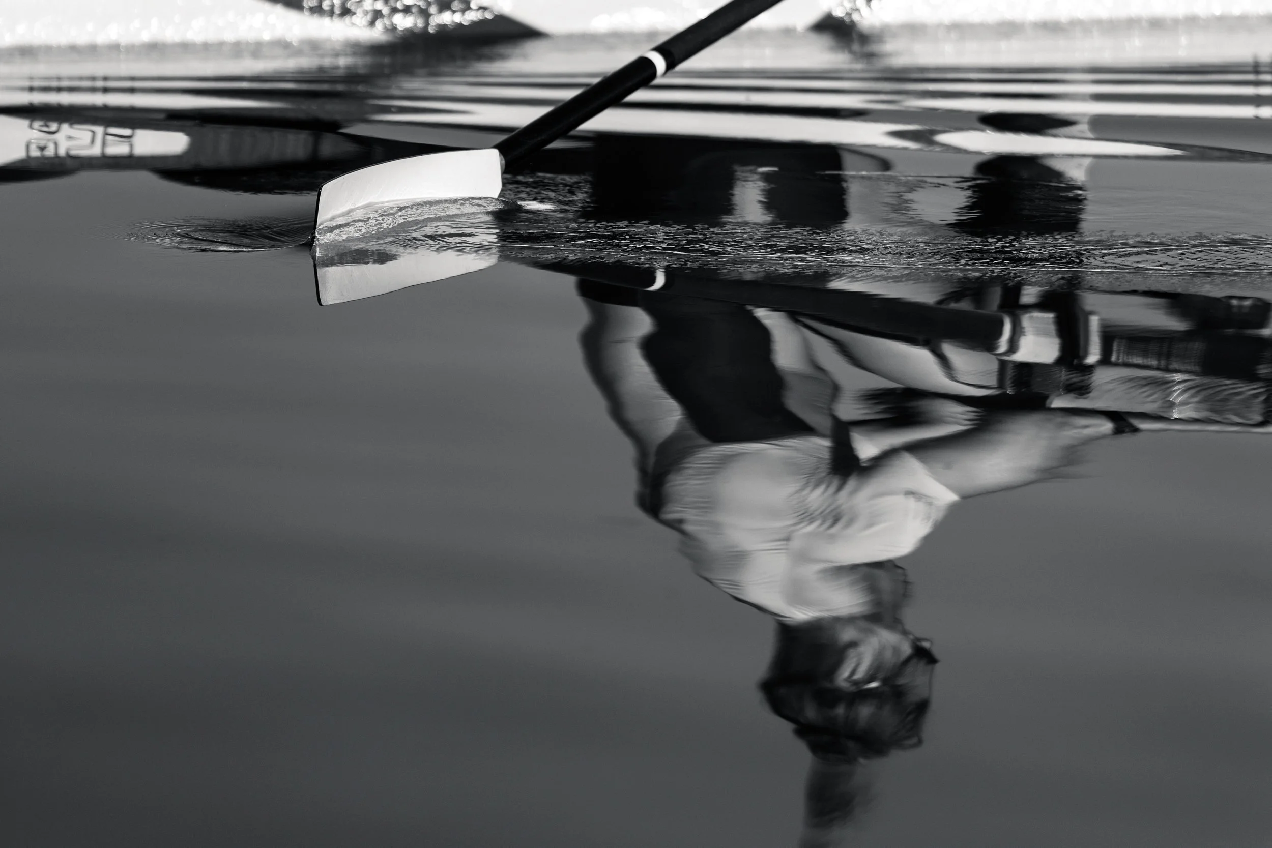 A person in a swimsuit stands on a kayak or paddleboard, reflected in the calm water beneath them, with a paddle resting on the water's surface.