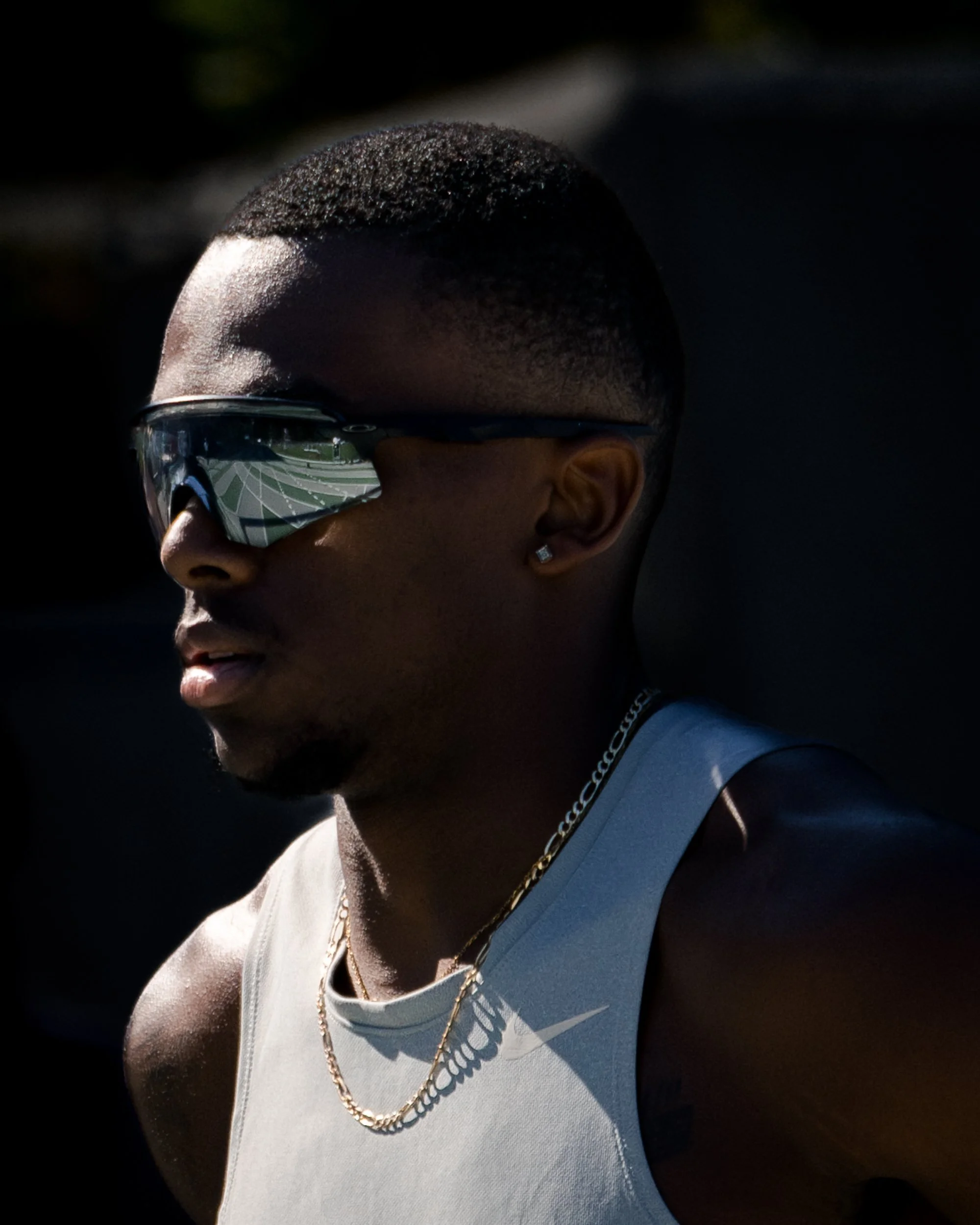 Close-up profile of a young man with dark skin wearing reflective sports sunglasses, a white tank top, gold chain, diamond earring, and sporting a short haircut, outdoors in bright sunlight.