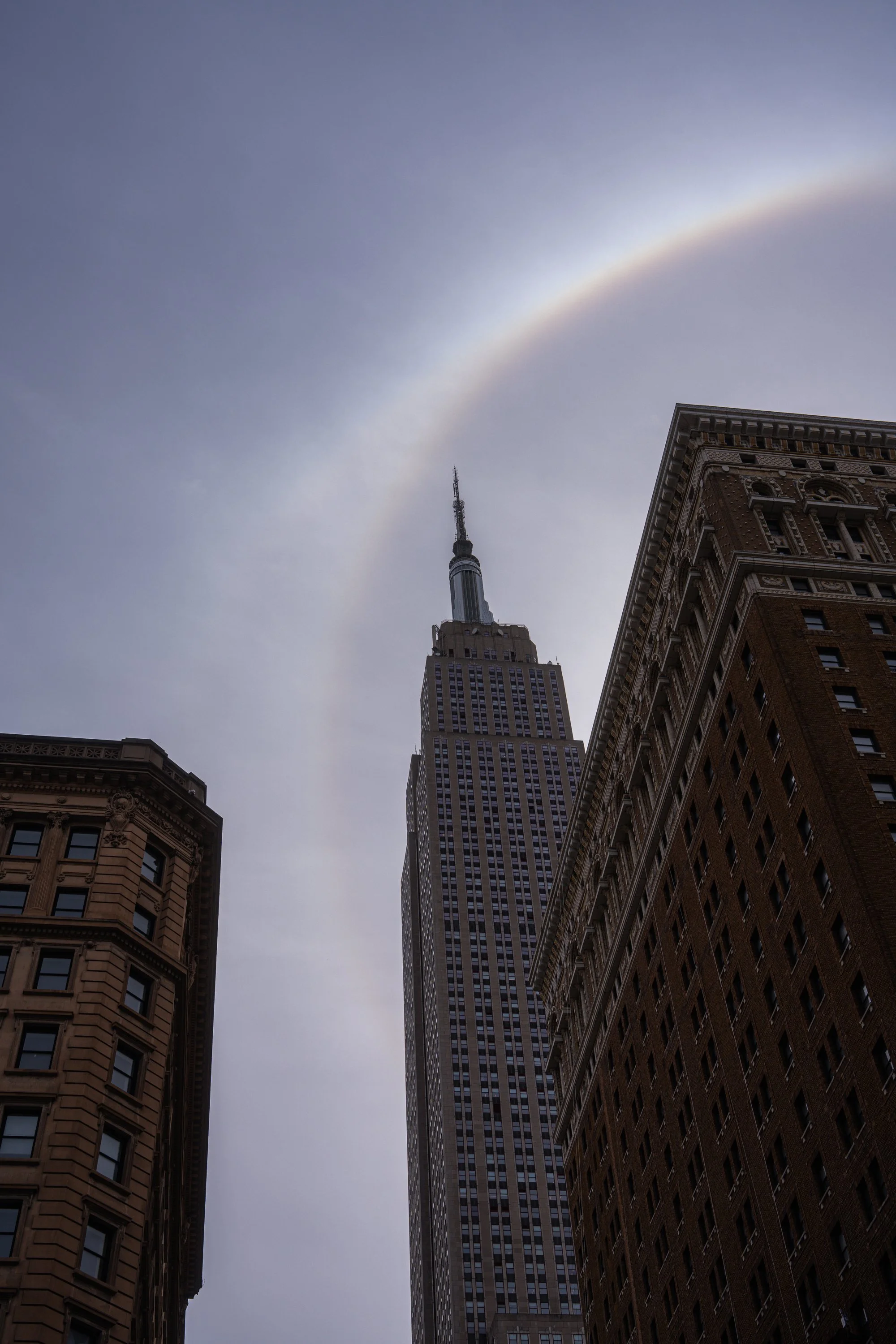 A cityscape featuring tall buildings with the Empire State Building in the center, under a cloudy sky with a faint rainbow arching above.