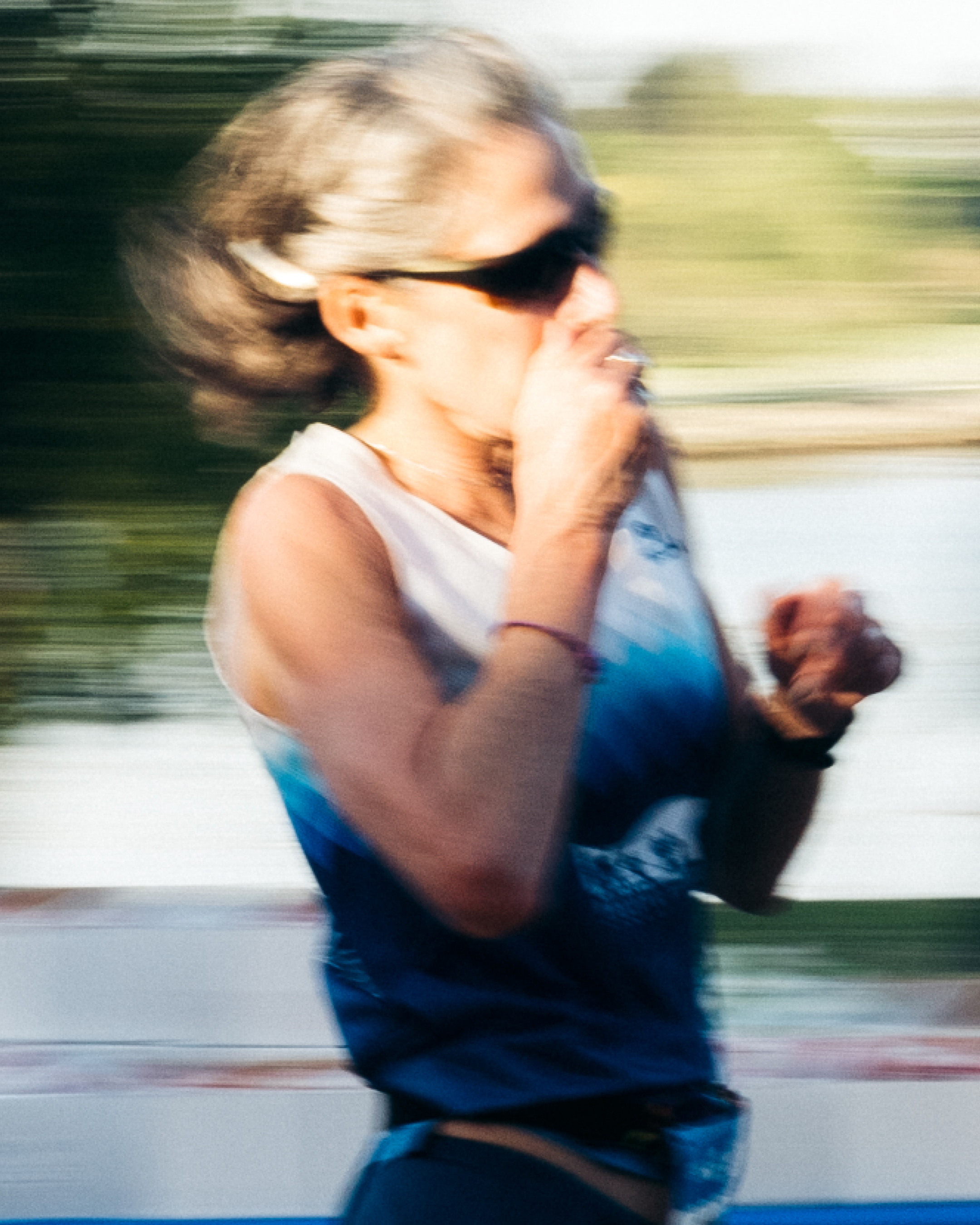 A woman with gray hair wearing sunglasses, a white sleeveless top, and a black glove, appears to be running or in motion outside, with a blurred background indicating speed.