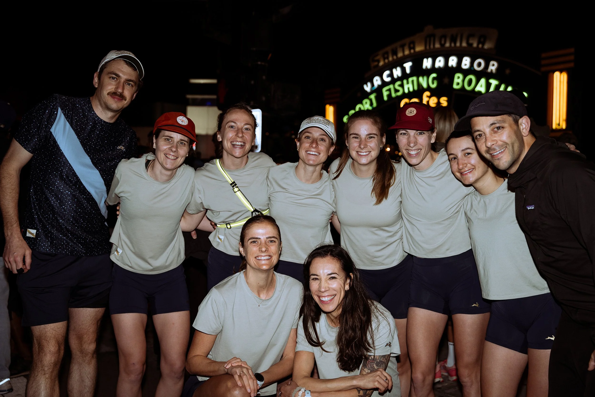 A group of smiling people, mostly women, posing together at night at an outdoor event near sign that reads 'Yacht Harbor Sport Fishing Boats'.