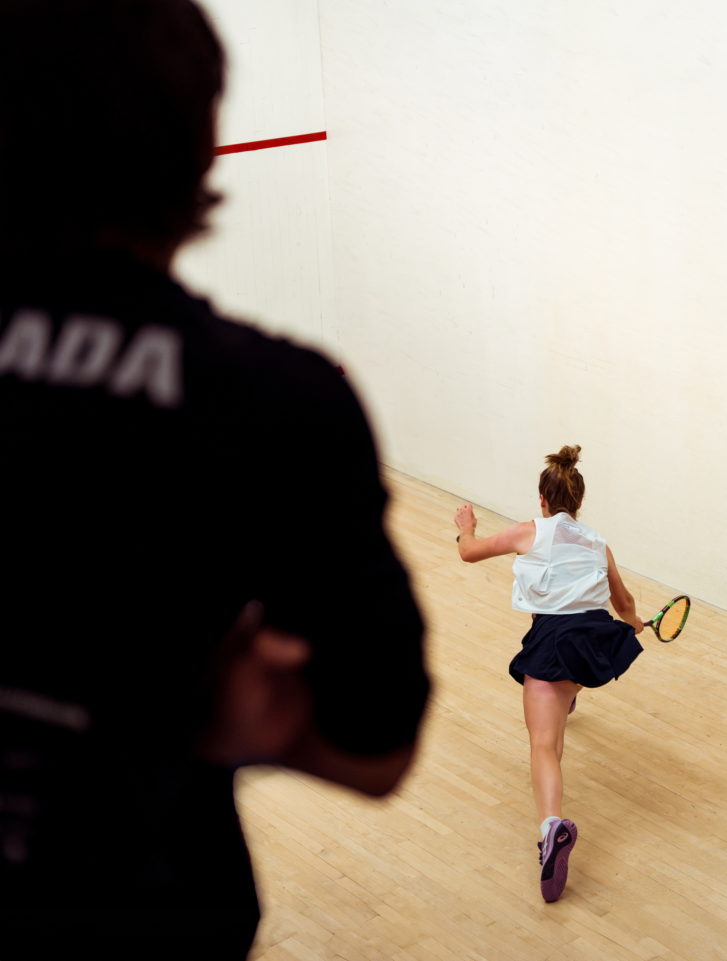 A woman playing squash on an indoor court, with another person watching in the foreground.