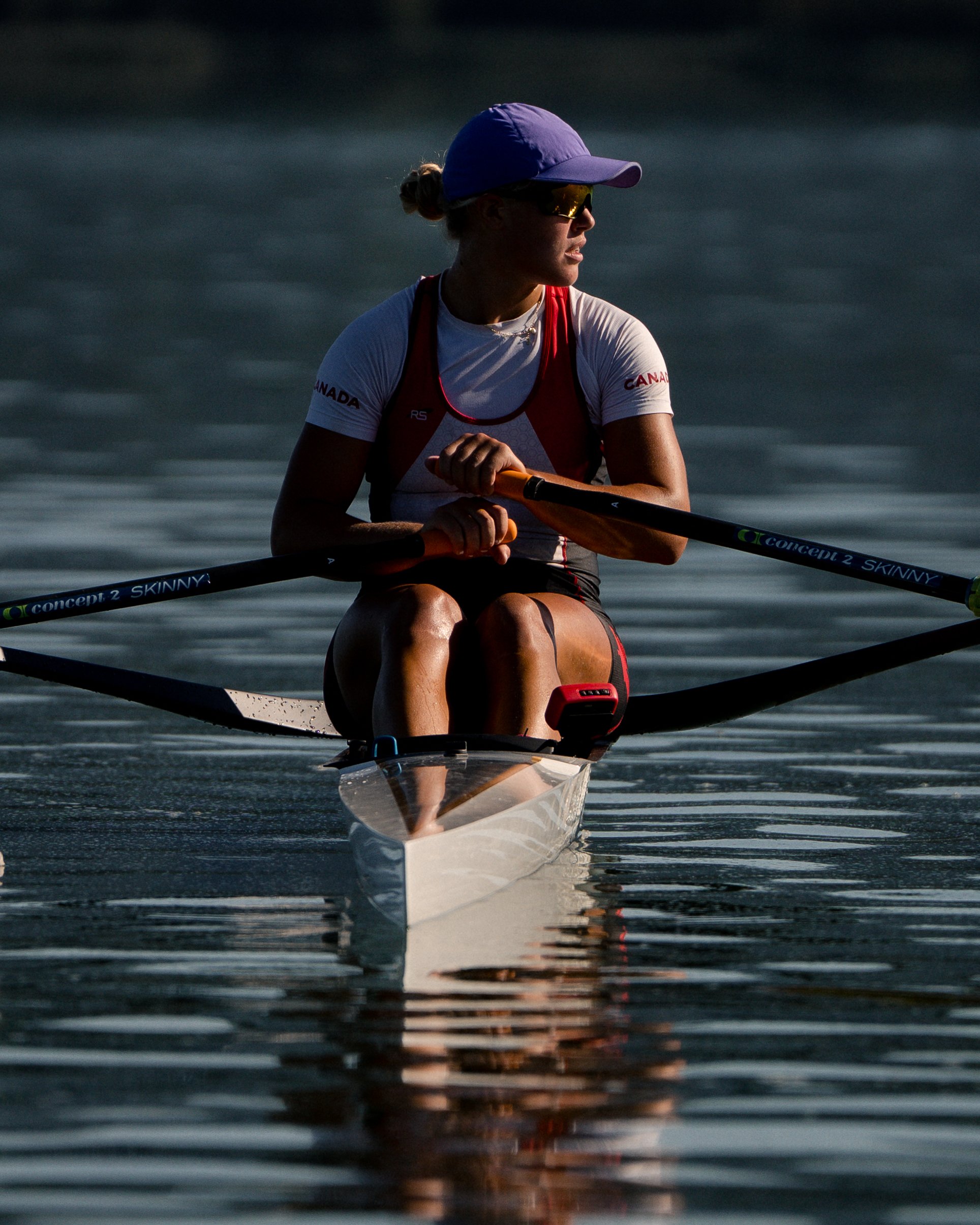 A woman kayaking on a calm body of water during late afternoon or early evening, wearing a purple cap, sunglasses, and athletic clothing with Canadian logos.