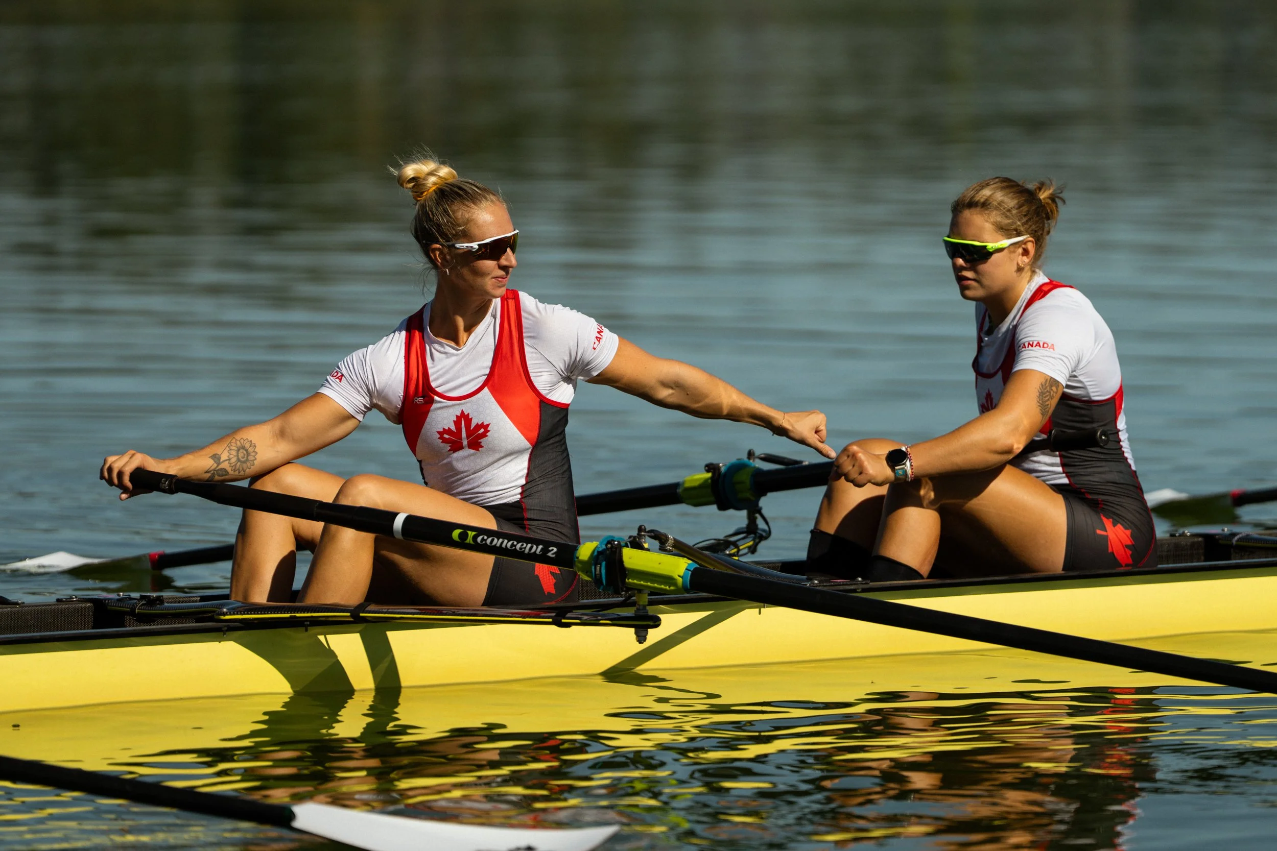 Two female Canadian rowers in a yellow boat on a calm lake, wearing matching red, black, and white athletic uniforms with Canadian maple leaf logos, sunglasses, and wristwatches, engaging in a rowing activity.