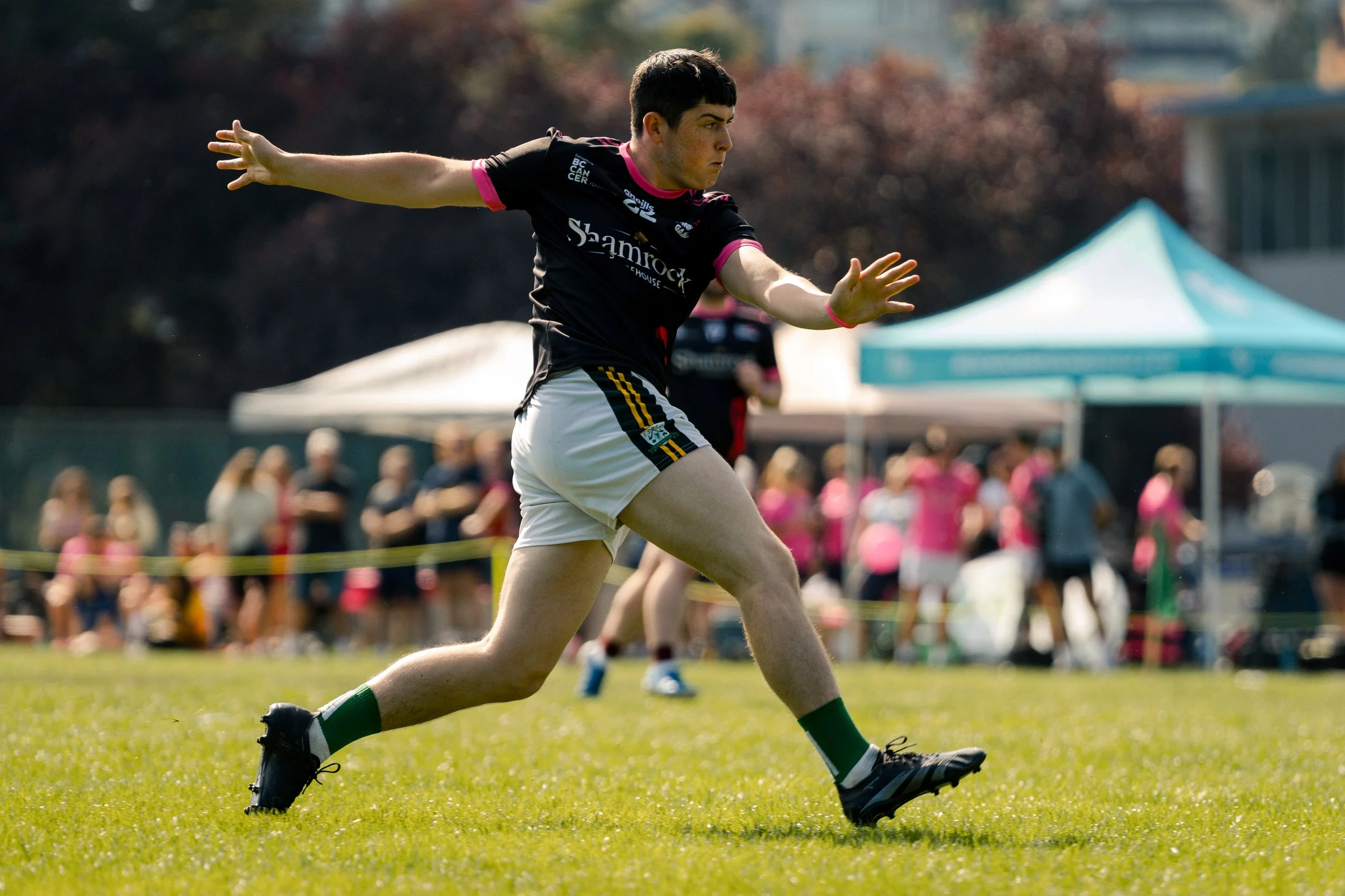 A young man playing rugby on a grassy field, wearing a black jersey with pink accents and white shorts, with a crowd and tents in the background.