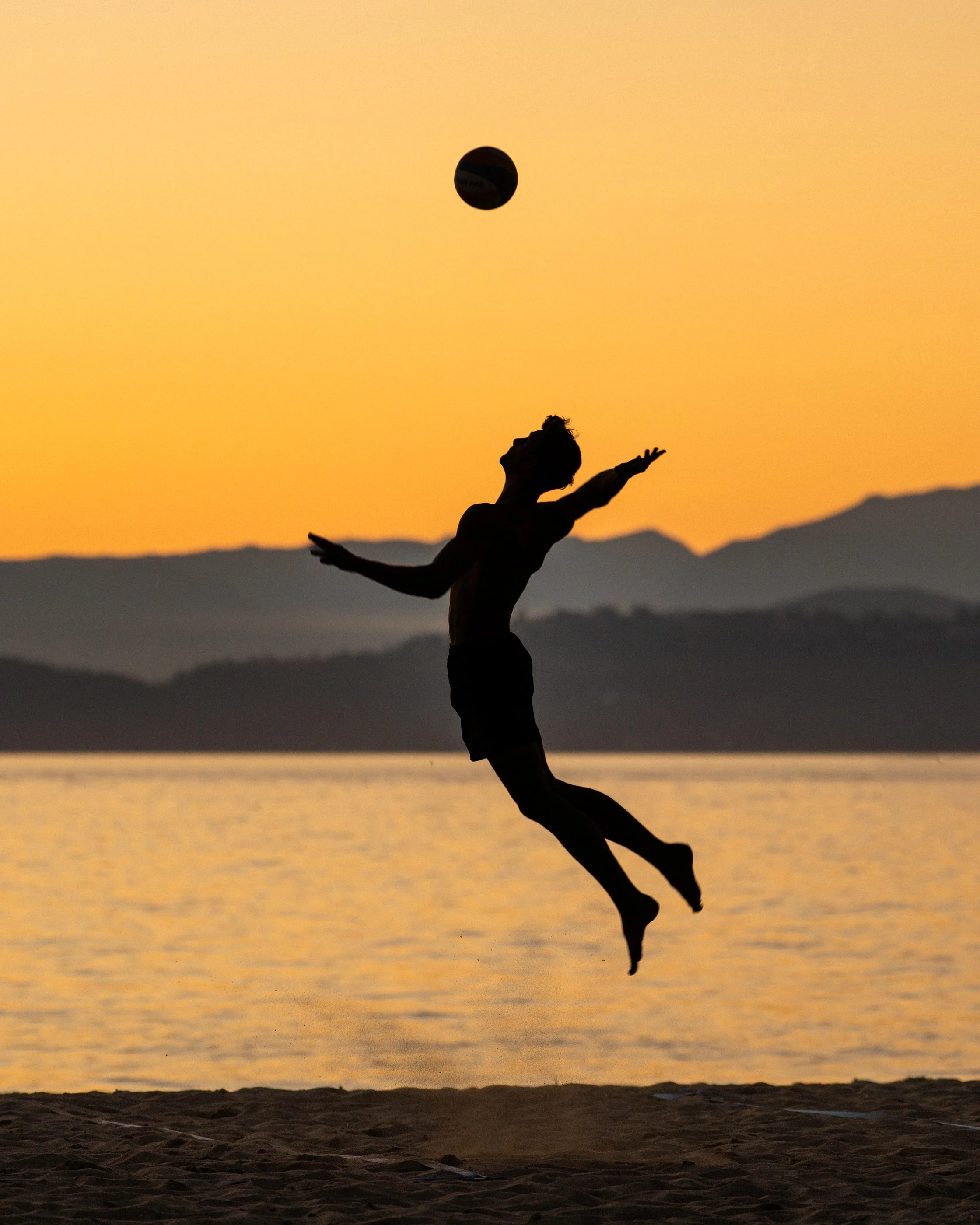 Silhouette of a person jumping at the beach during sunset, with a ball in the air and mountainous landscape in the background.