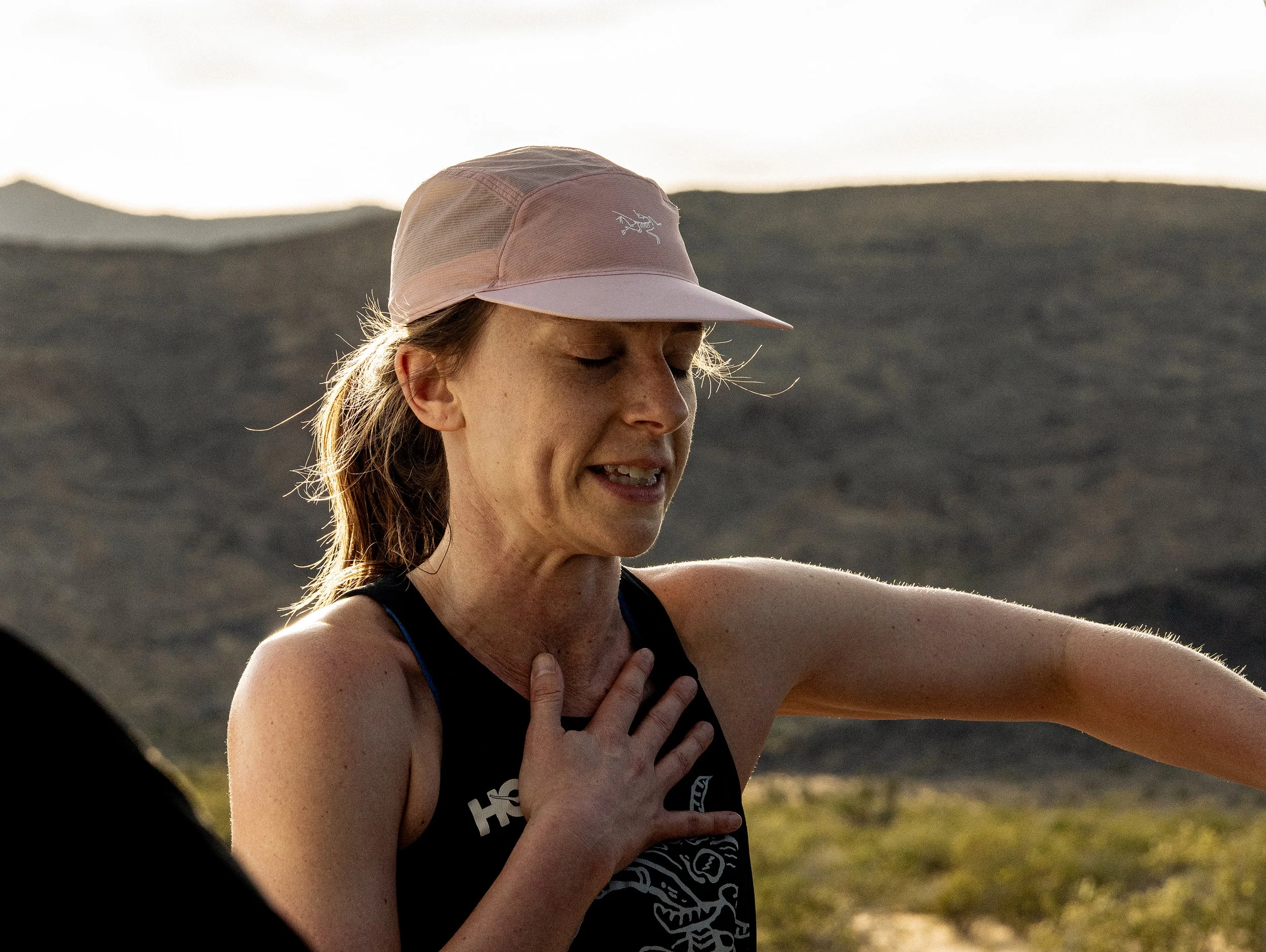 A woman wearing a pink cap and a black athletic top with a skeleton design, outdoors near mountains during sunset.
