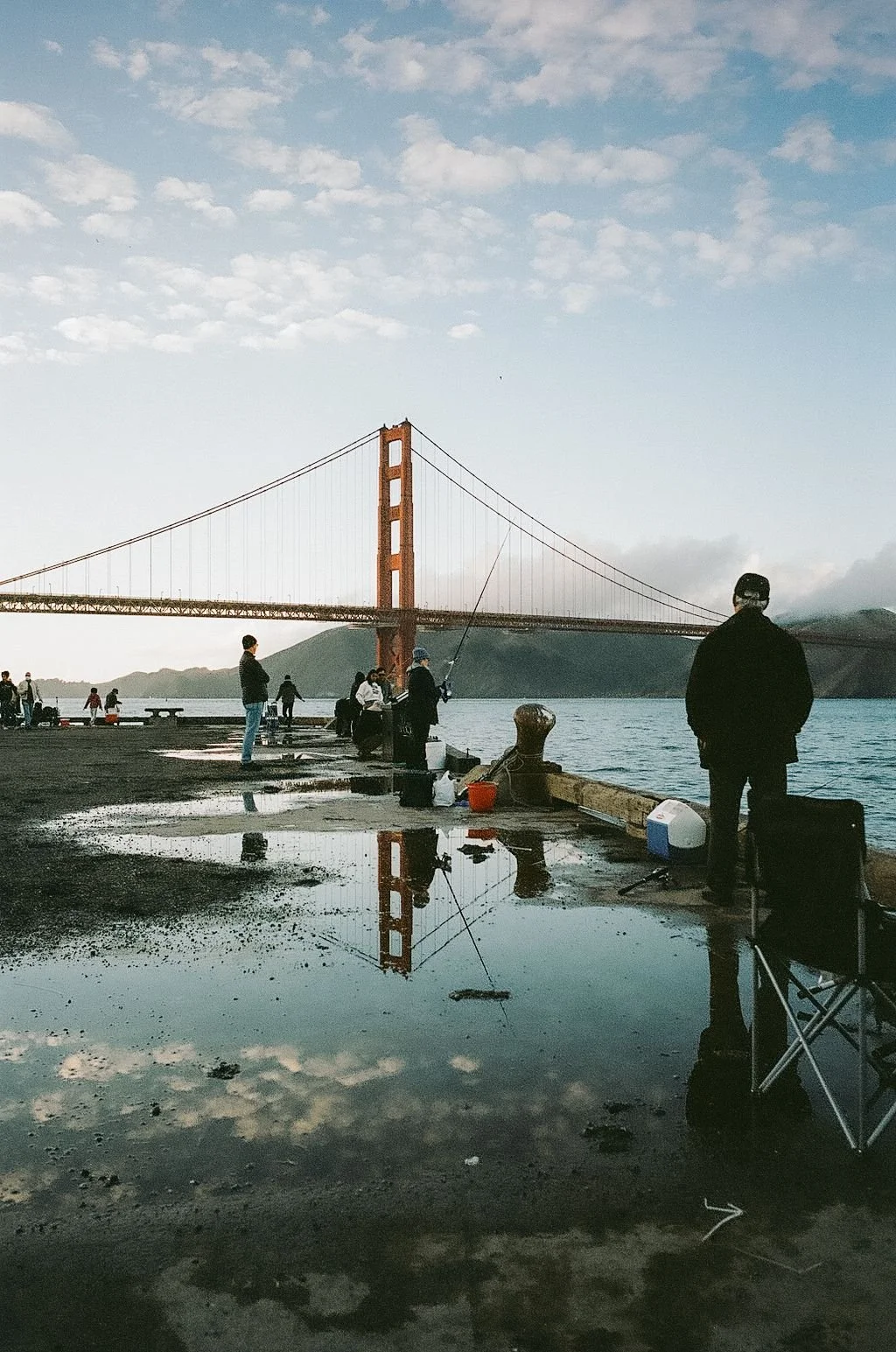 People fishing at a pier near the Golden Gate Bridge, with puddles reflecting the sky and bridge in San Francisco.