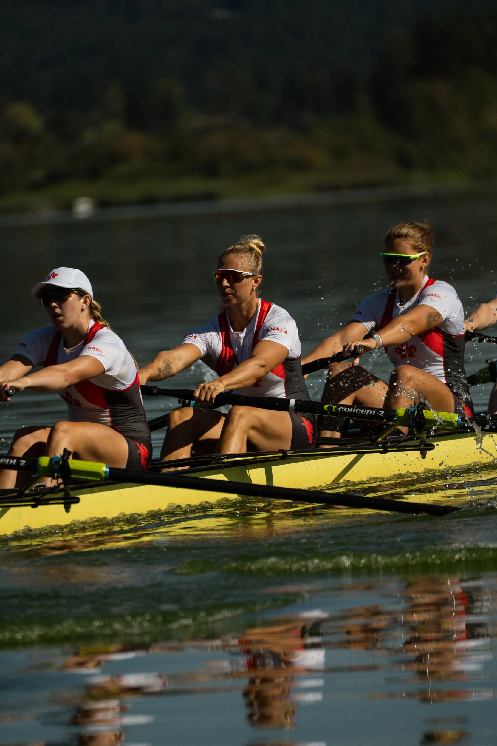 Three women wearing sunglasses and athletic clothing rowing a yellow boat on a lake.