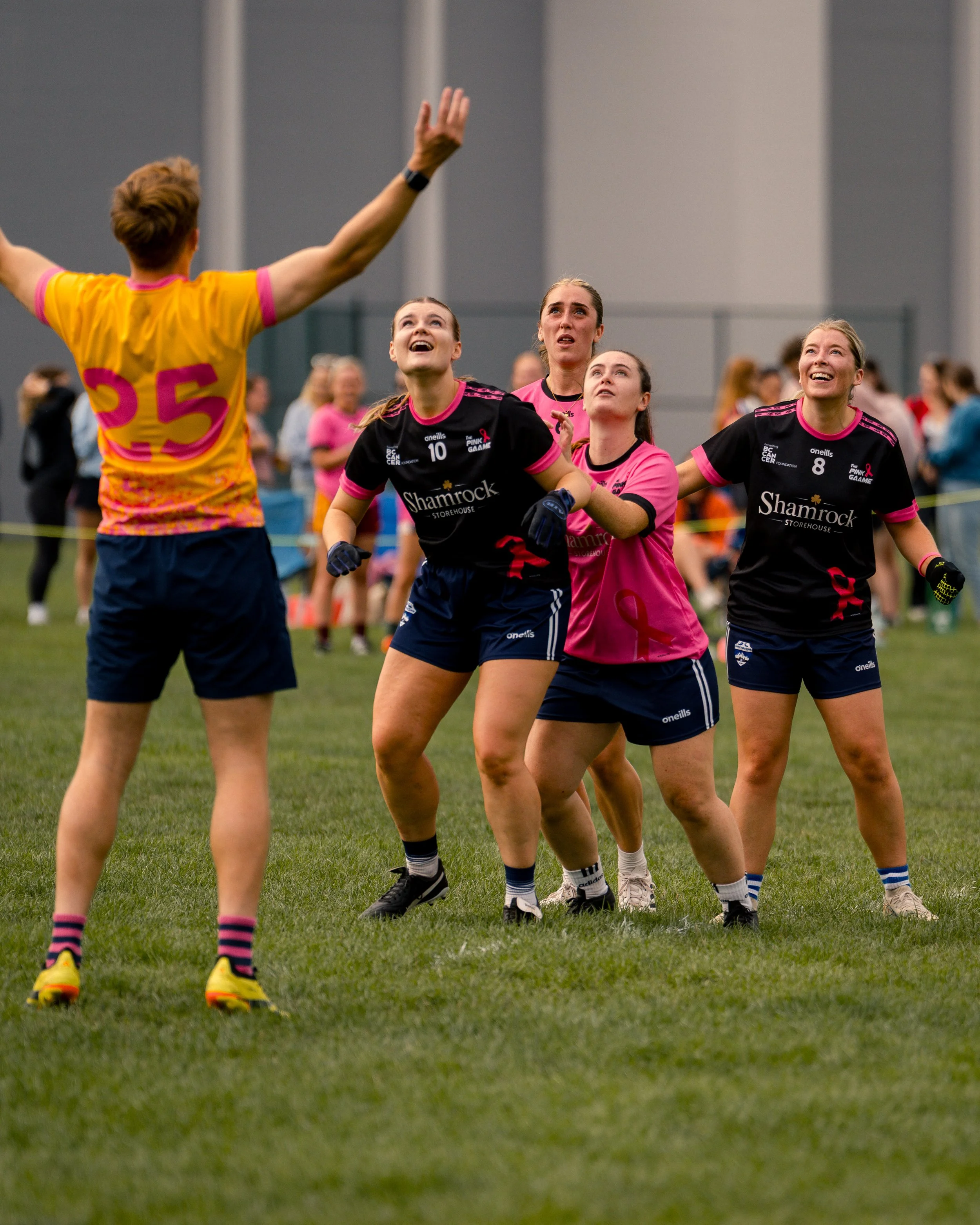 A group of female rugby players on the field during a game, with one player in a yellow jersey facing away and the others in black and pink jerseys looking upward.