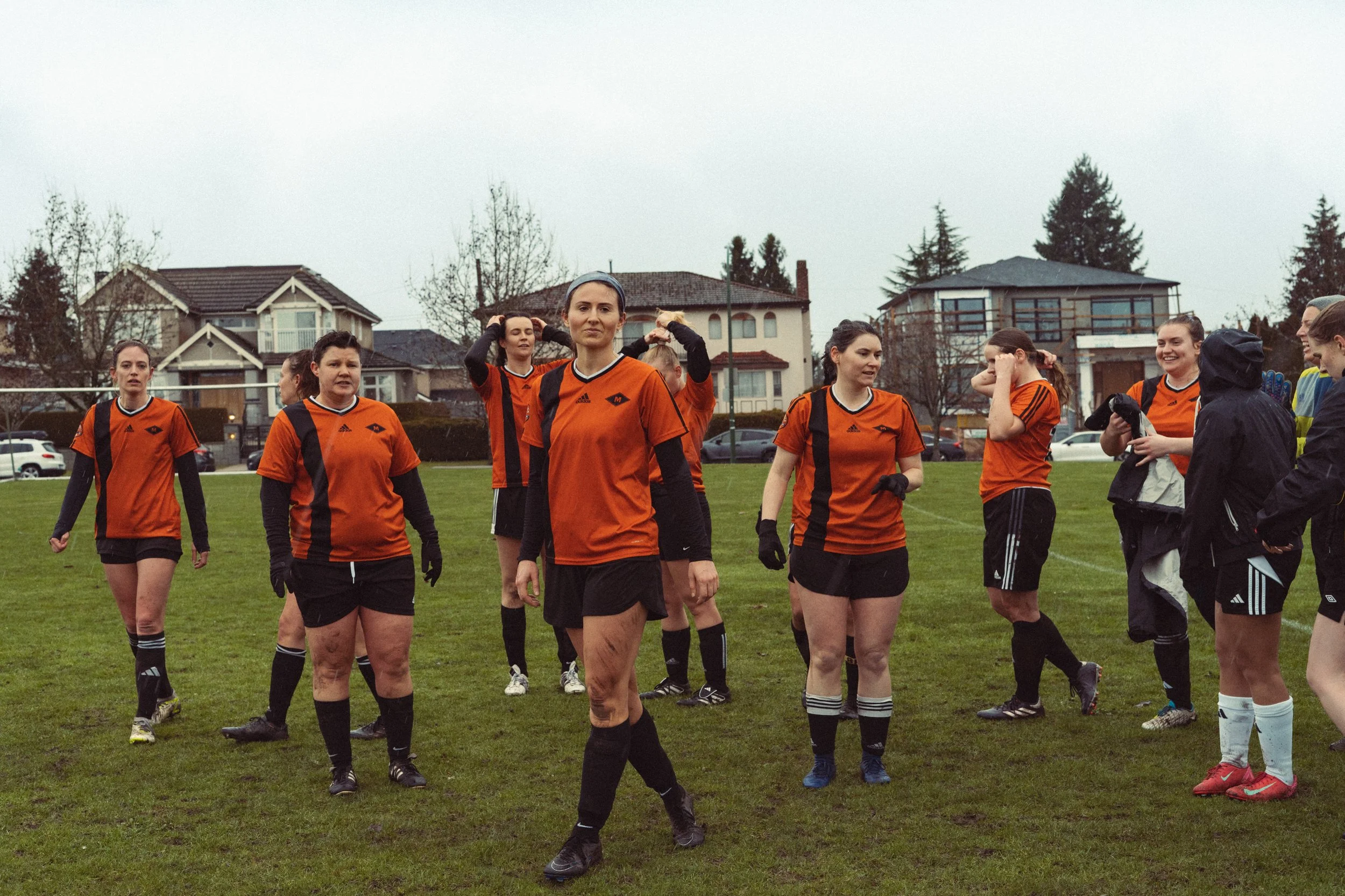 A group of women soccer players wearing orange jerseys and black shorts on a grassy field, some adjusting their hair, with houses and trees in the background on a cloudy day.
