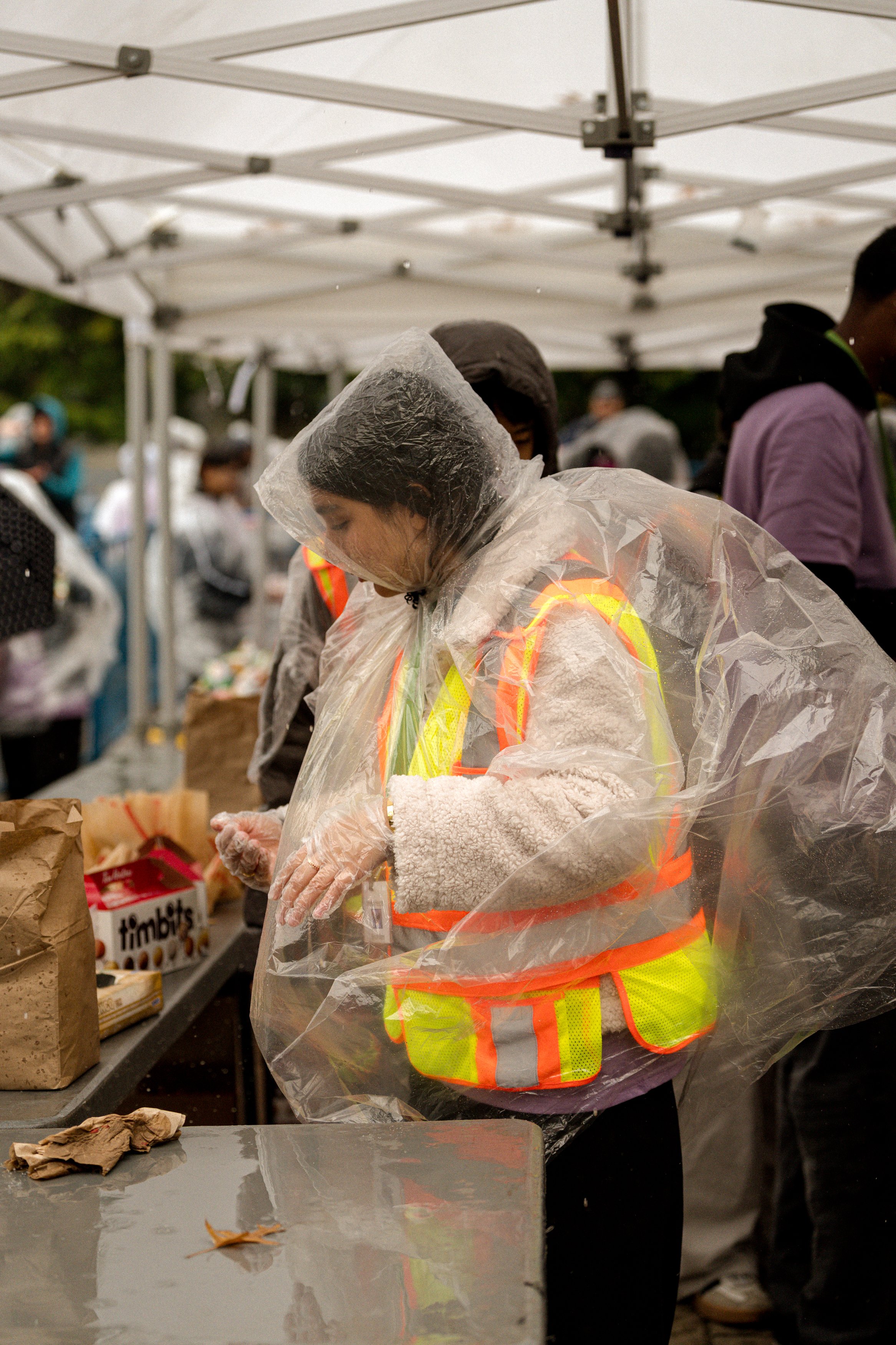 A person wearing a clear rain poncho and a reflective safety vest at an outdoor market or event, with other people and tents visible in the background.