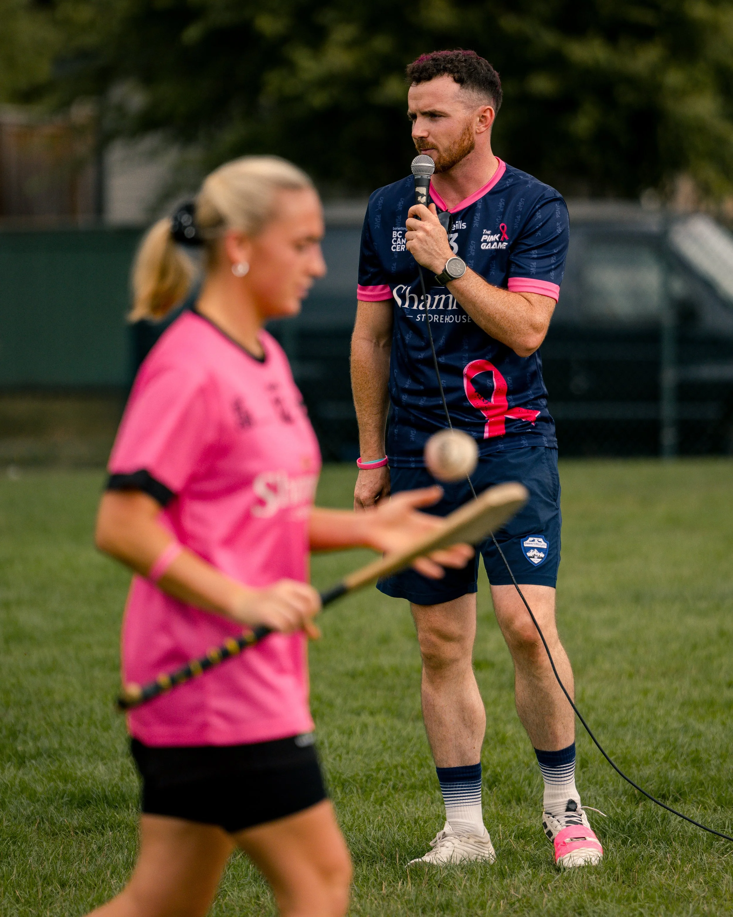 A man with short curly hair and a beard wearing a pink and navy sports jersey, navy shorts, white socks with pink stripes, and pink sneakers, holding a microphone on a grassy field. A woman in a pink sports jersey and black shorts, holding a paddle, 