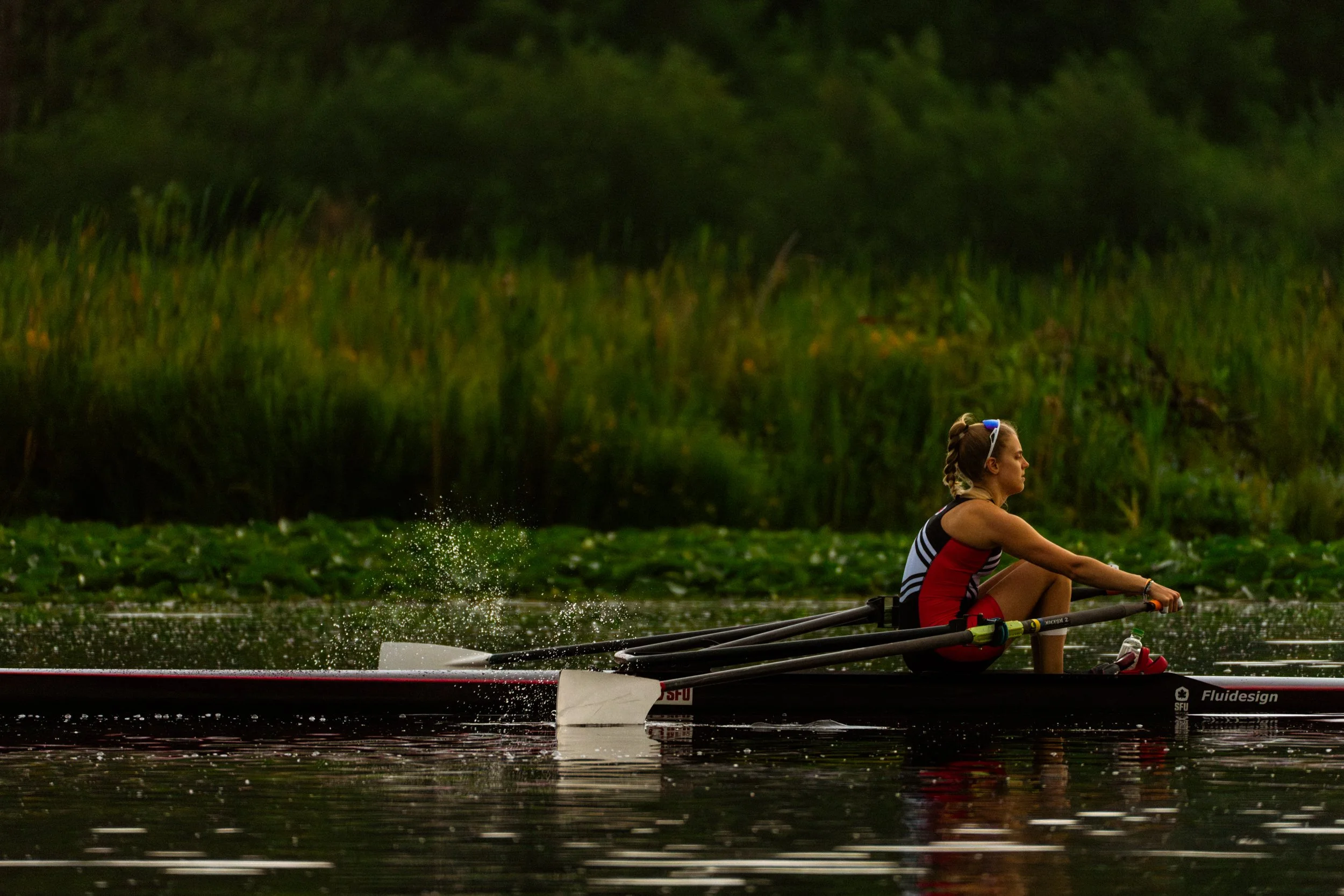 A woman rowing a boat on a calm body of water with green reeds and trees in the background.