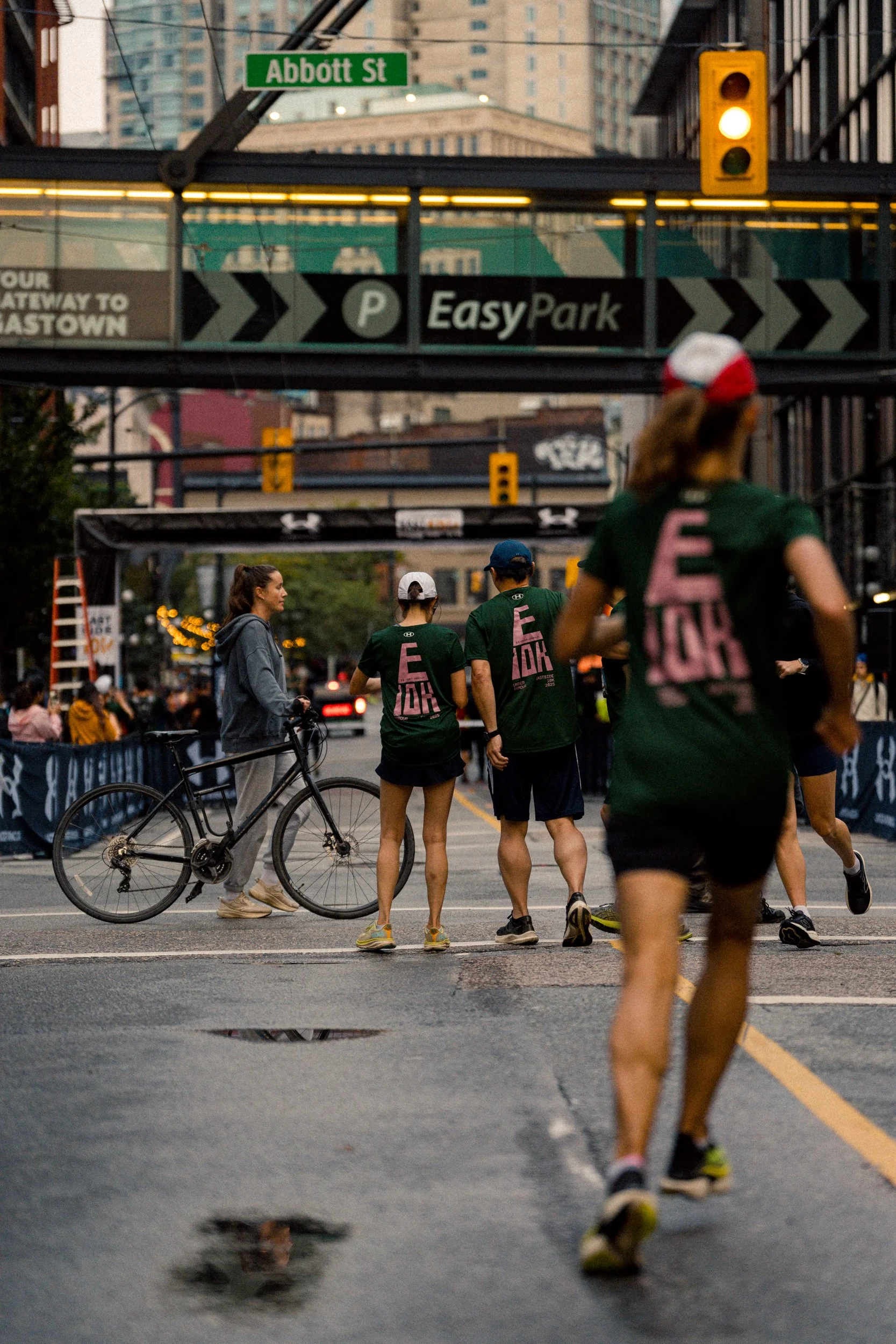People participating in an outdoor running event on a city street, with some wearing matching shirts and one with a bicycle, under an overhead walkway with signs for EasyPark and Abbott Street, in an urban environment.