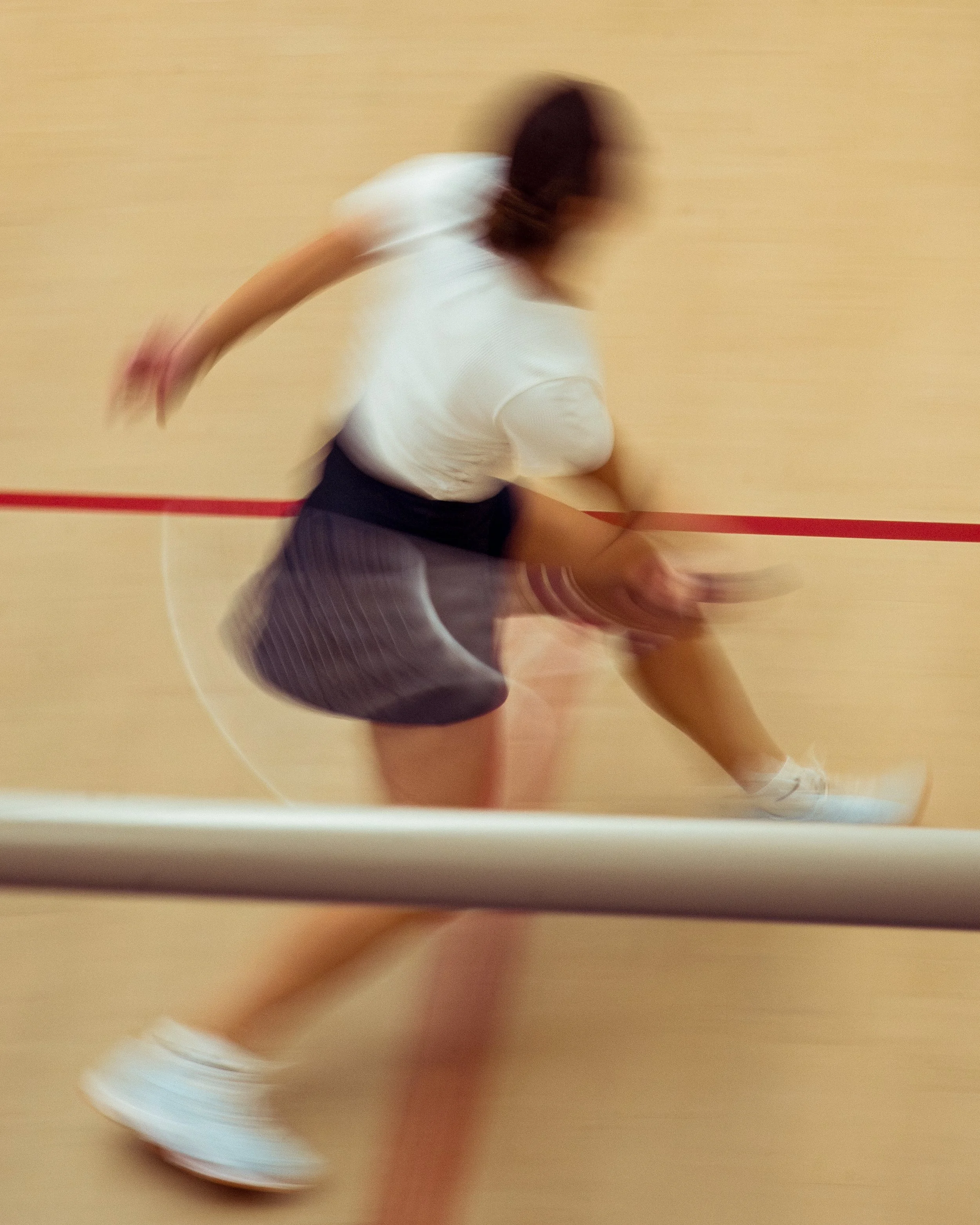 Blurred image of a person running indoors on a gym or sports court, wearing a white shirt, striped shorts, and white sneakers, with a red line on the floor in the background.