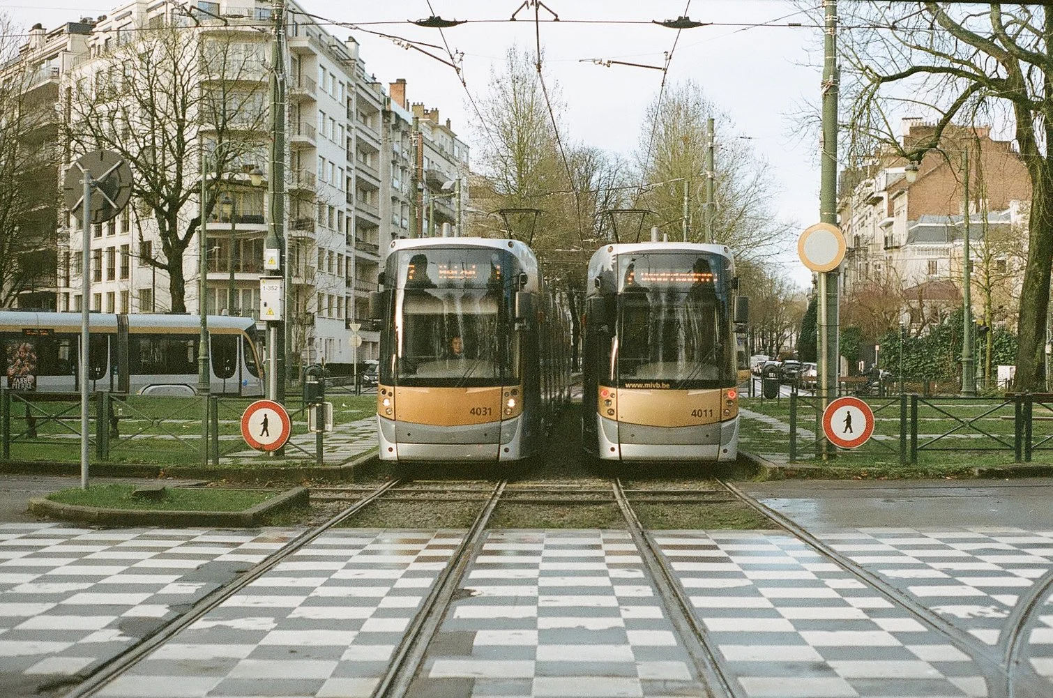 Two modern streetcars with beige and black coloring on tracks in an urban area, with city buildings, trees, and other vehicles in the background.
