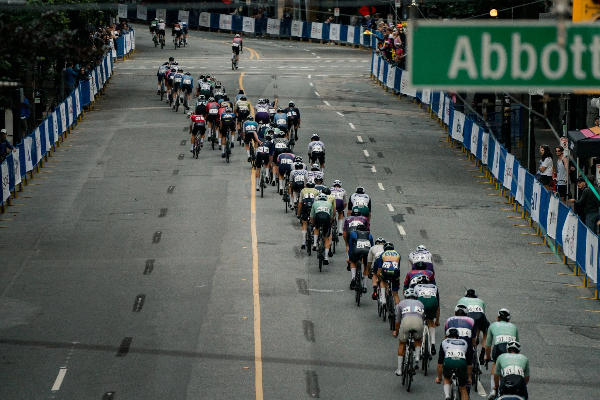 A group of cyclists participating in a race on a city street, with spectators watching from the sides and barriers lining the road.