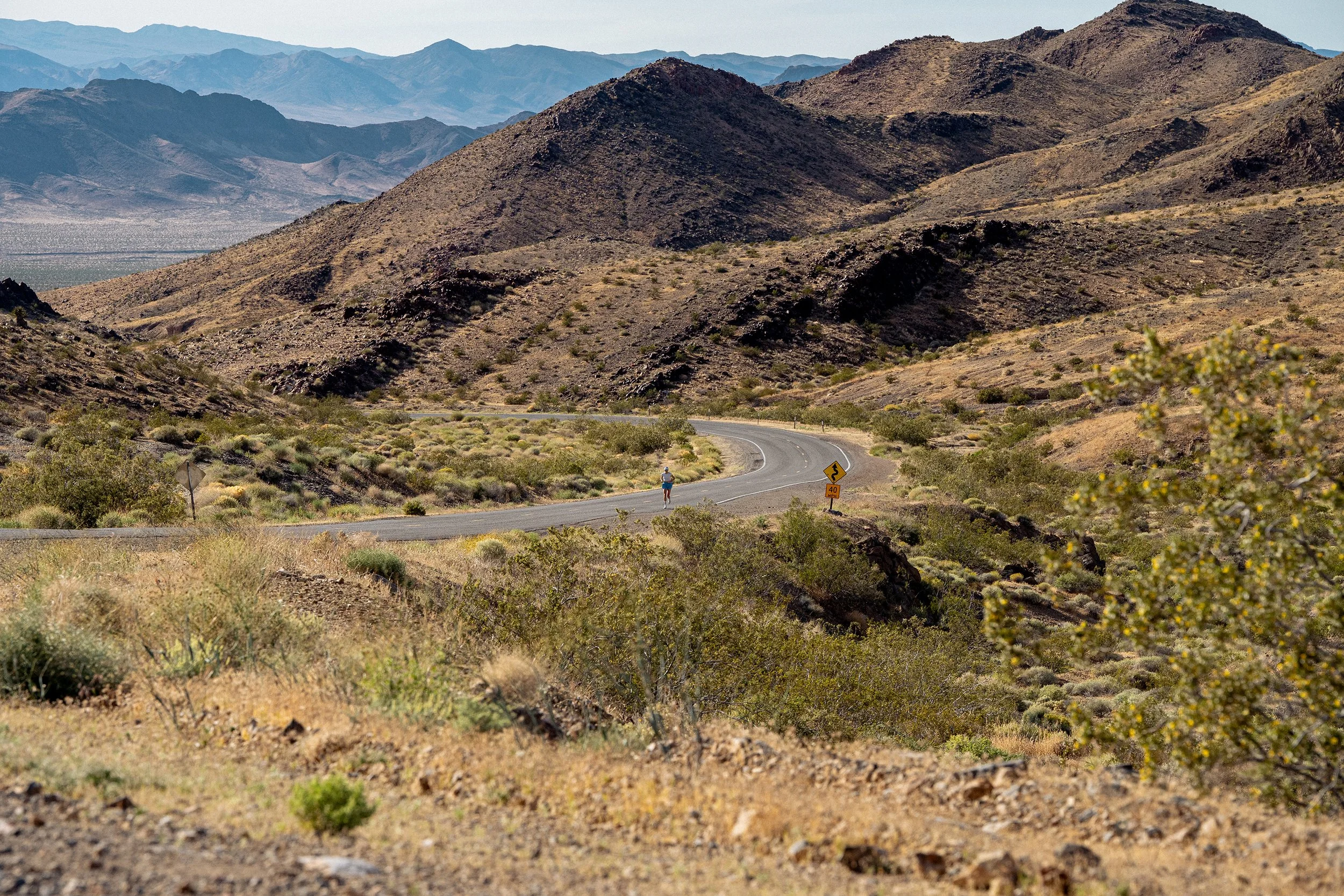 A lone person walking on a winding road through a desert landscape with brown, rocky hills and mountains in the background under a clear sky.