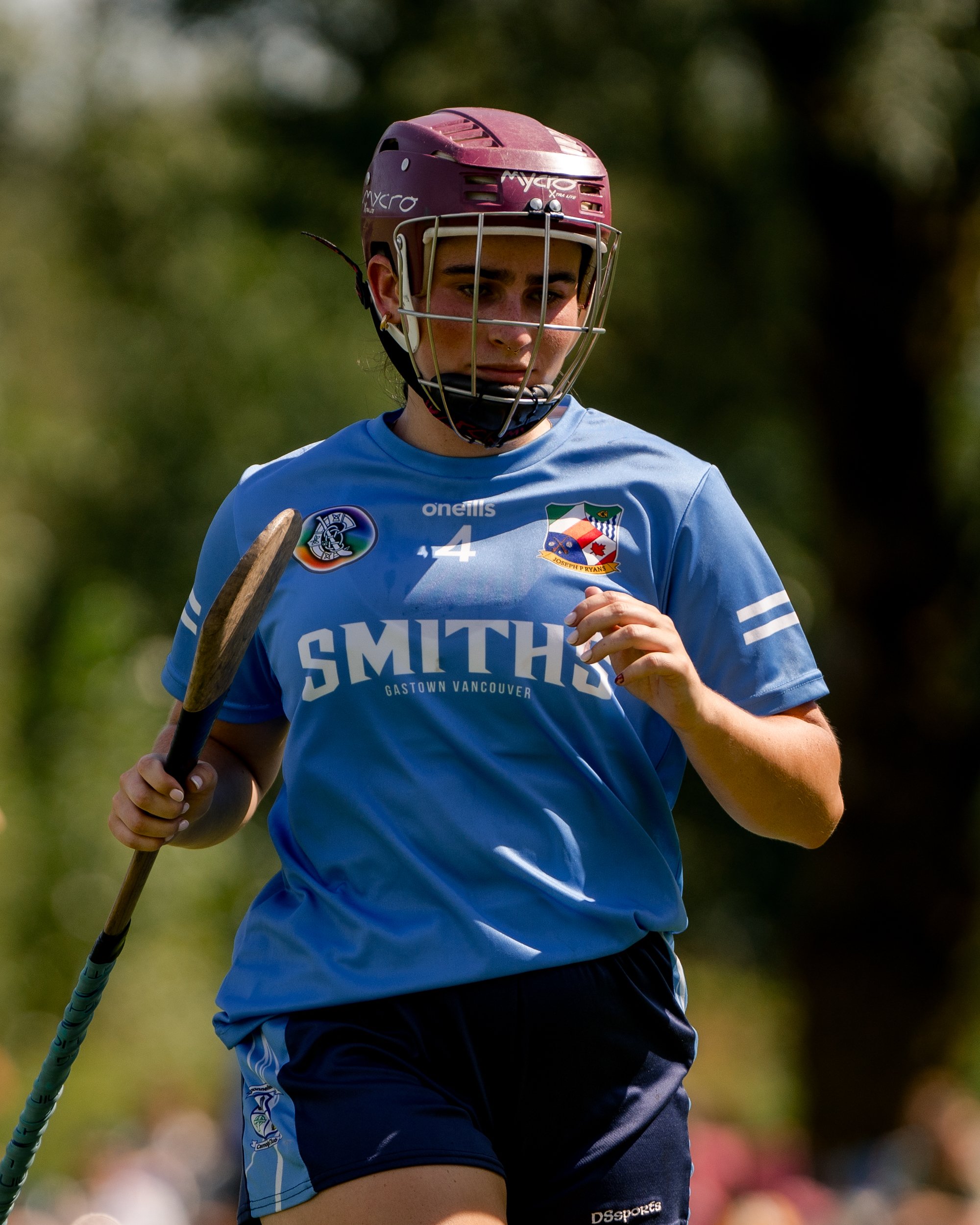 A female lacrosse player wearing a maroon helmet and a sky blue jersey with the word 'SMITHS' and the location 'Gastown Vancouver' on it, holding a lacrosse stick.