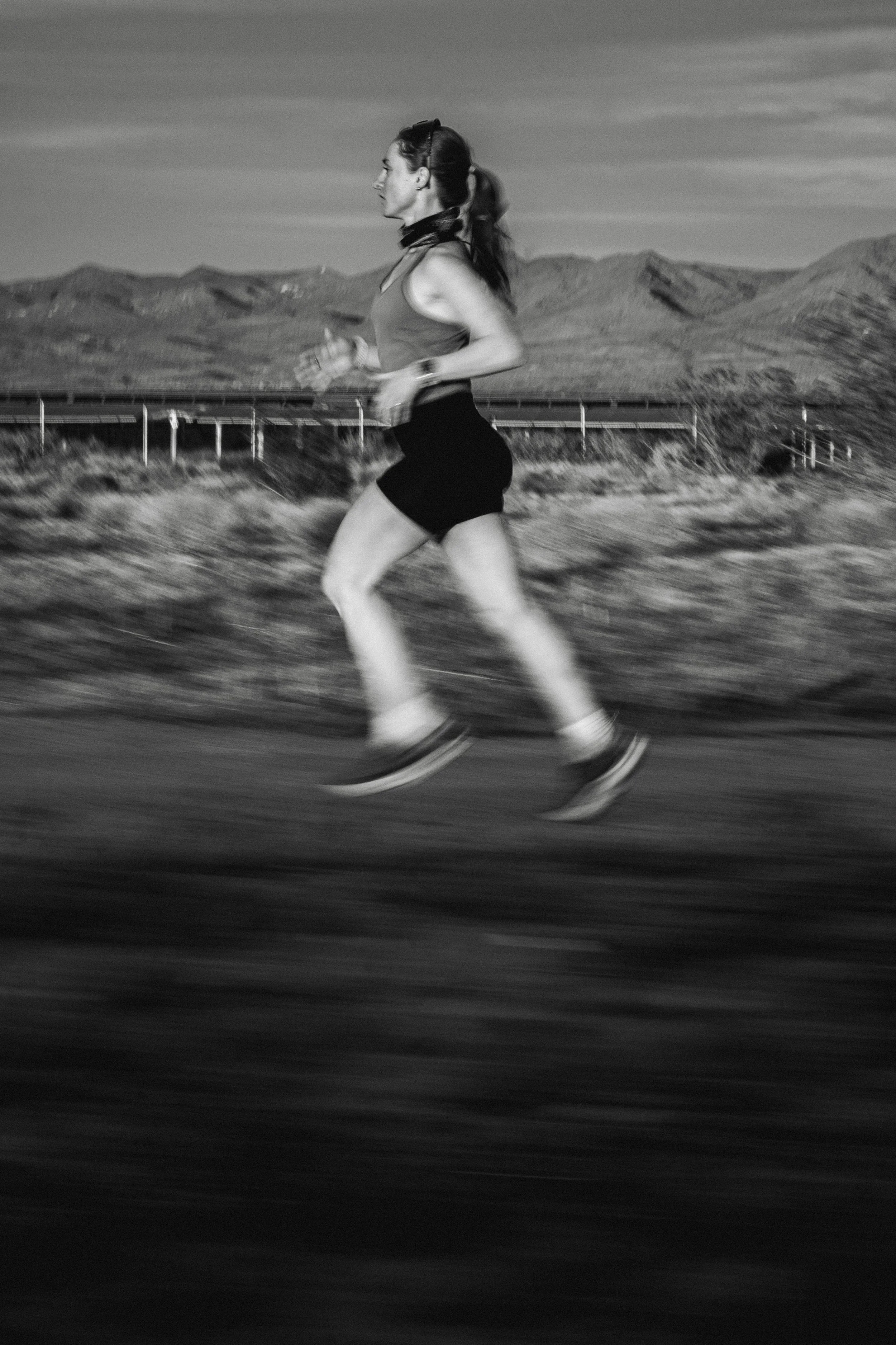 A woman jogging outdoors on a paved road with mountains and a clear sky in the background.