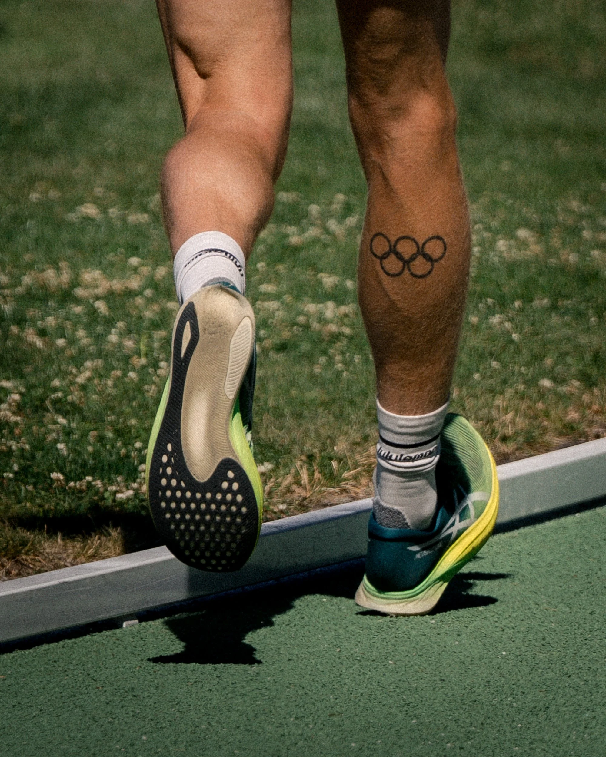 Close-up of a runner's legs and feet, one foot in mid-air, on a running track. The runner is wearing black, green, and white running shoes and white socks with black branding. A tattoo of five interlinked black circles is visible on the lower part of