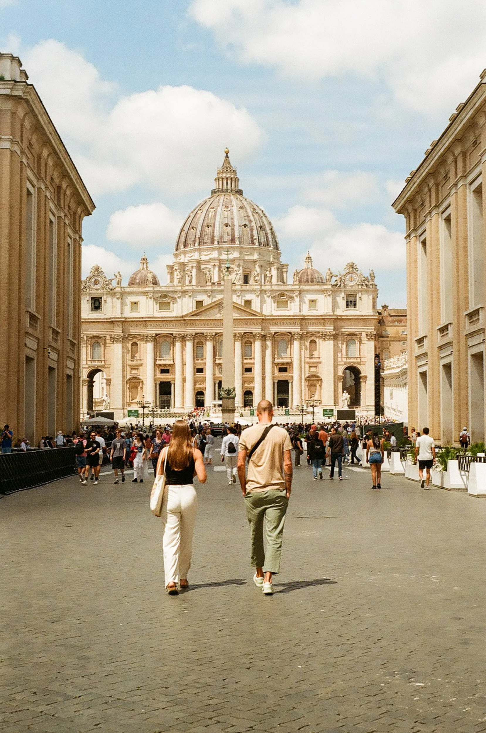 People walking towards St. Peter's Basilica in Vatican City, with a crowd gathered in the square and two women with beige pants and a man with green pants in the foreground.
