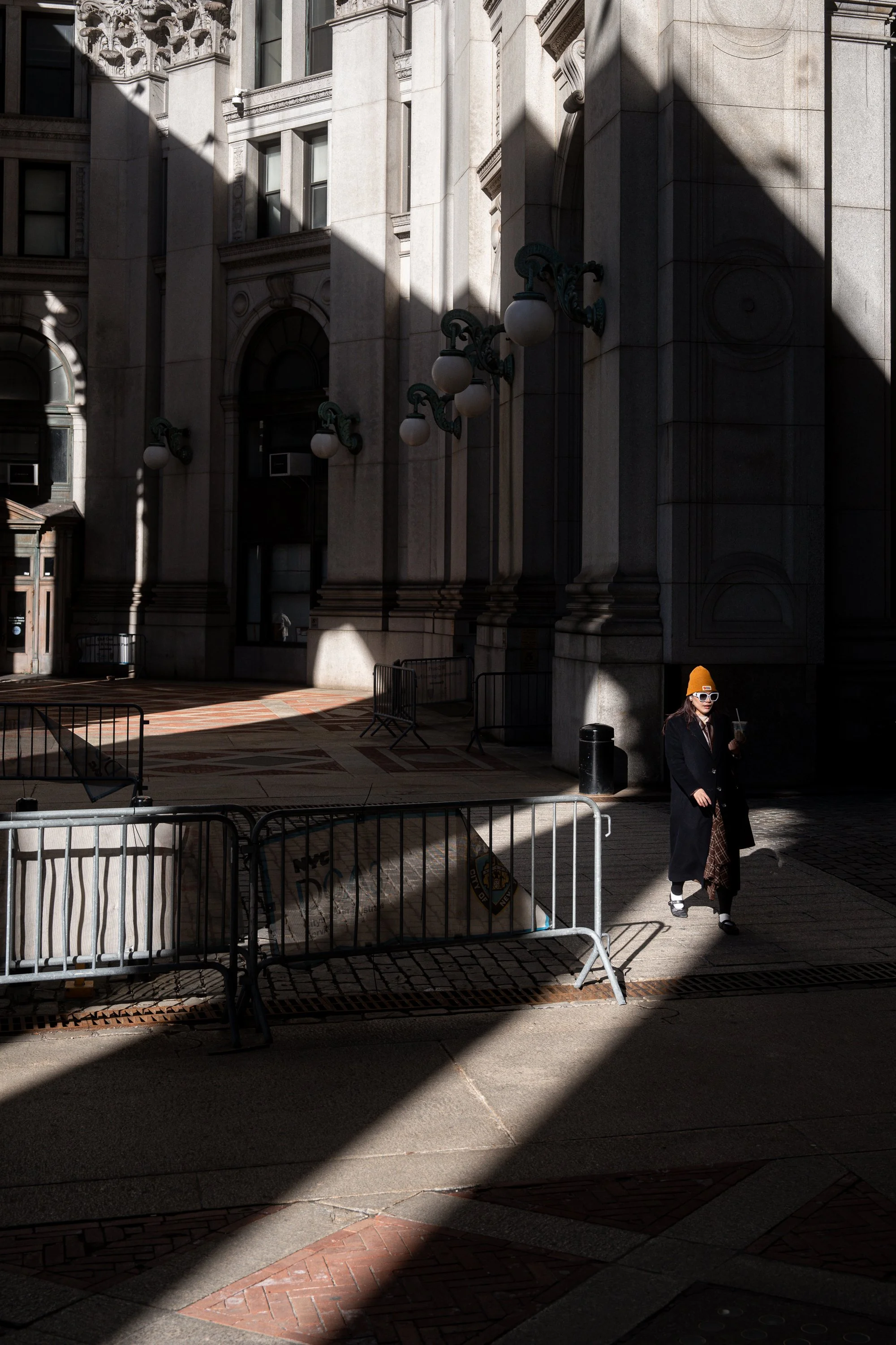 A woman wearing a yellow beanie, sunglasses, and a long coat walking on a city sidewalk, with a large stone building in the background and shadows cast across the scene.