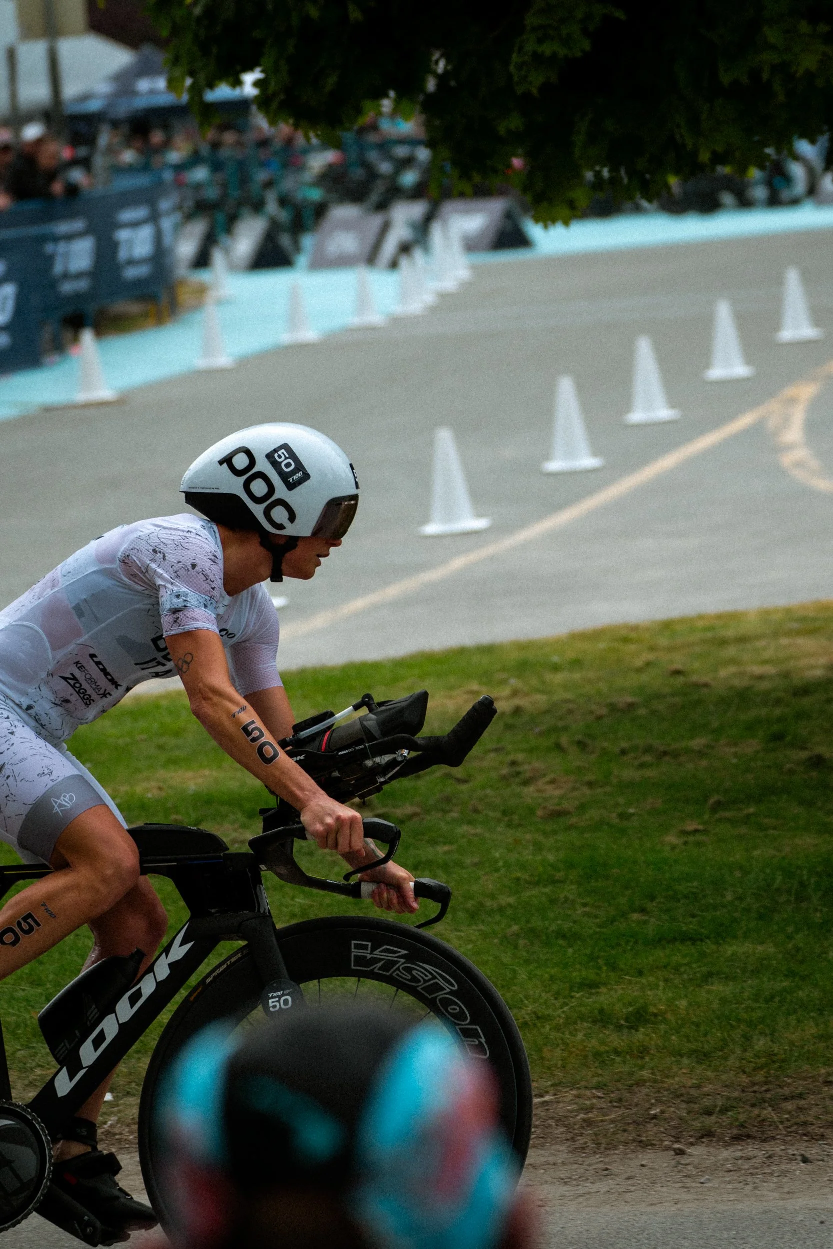 A cyclist in a white helmet and matching athletic outfit rides a black Dura-Ace time trial bike on a curved road, with a grassy area on the side and traffic cones marking the course, during a sporting event.