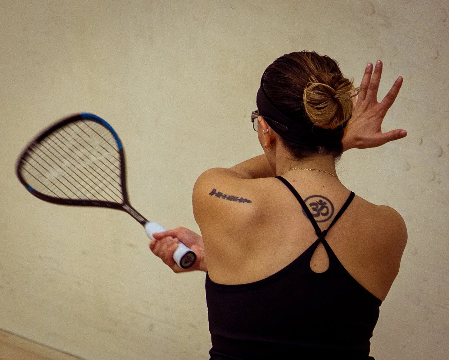 A woman with tattoos on her back and shoulder, wearing a black tank top and glasses, playing indoor squash, holding a racket in her right hand and touching the wall with her left hand.