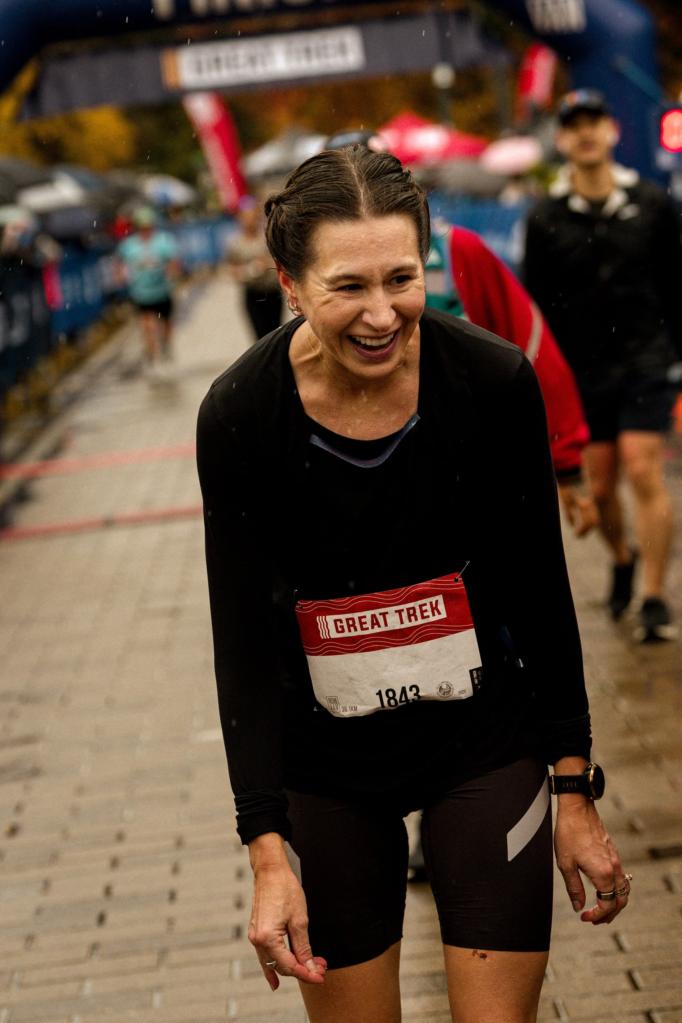 A woman smiling and laughing during a race, wearing a black shirt and shorts, with a racial bib number 1843 that says "Great Trek" on it. It is rainy with other runners and spectators in the background.