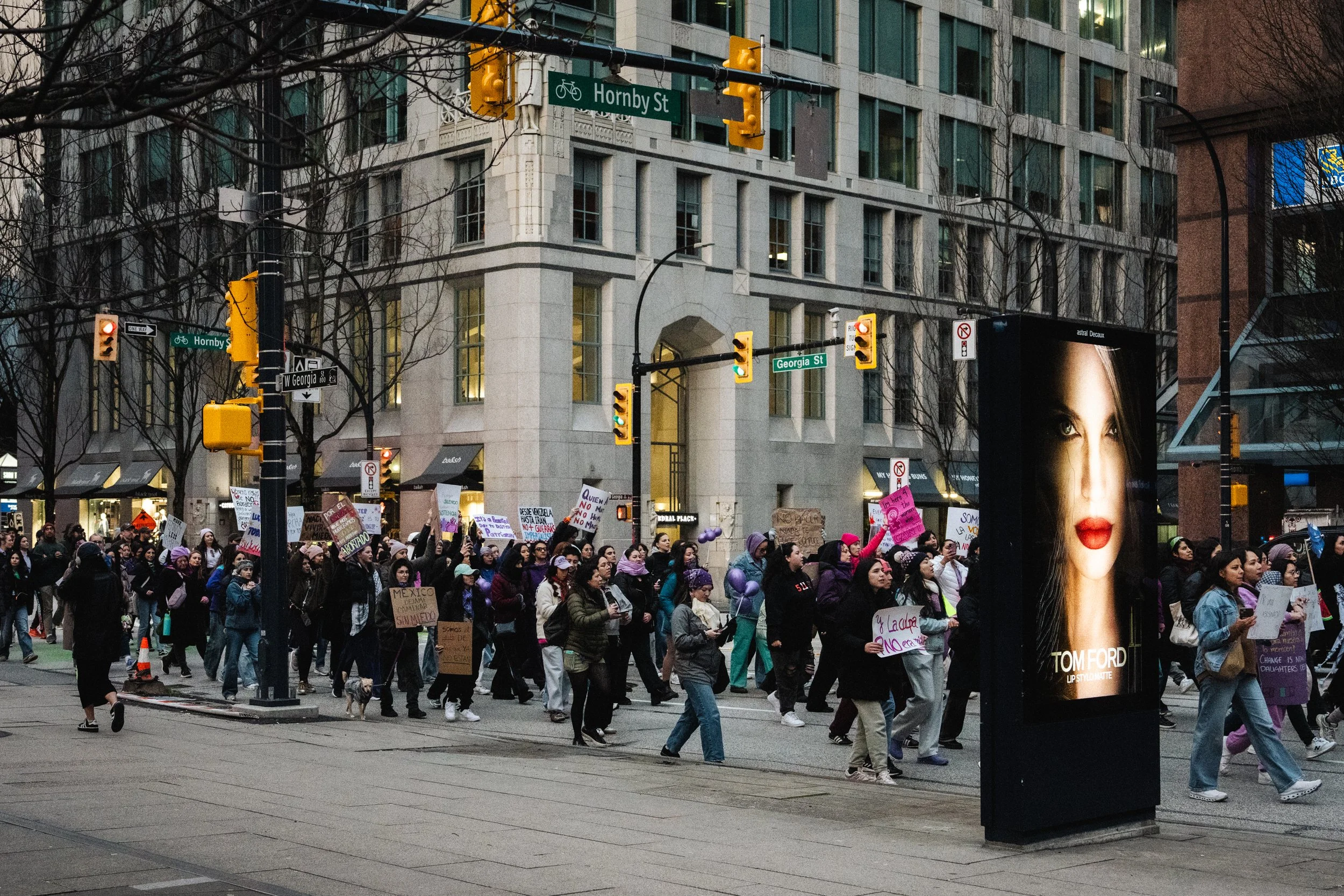 A crowd of protesters marching in an urban area at dusk, with banners and signs, a large advertisement featuring a woman's face, and city buildings with traffic lights and street signs for Hornby Street and Georgia Street.