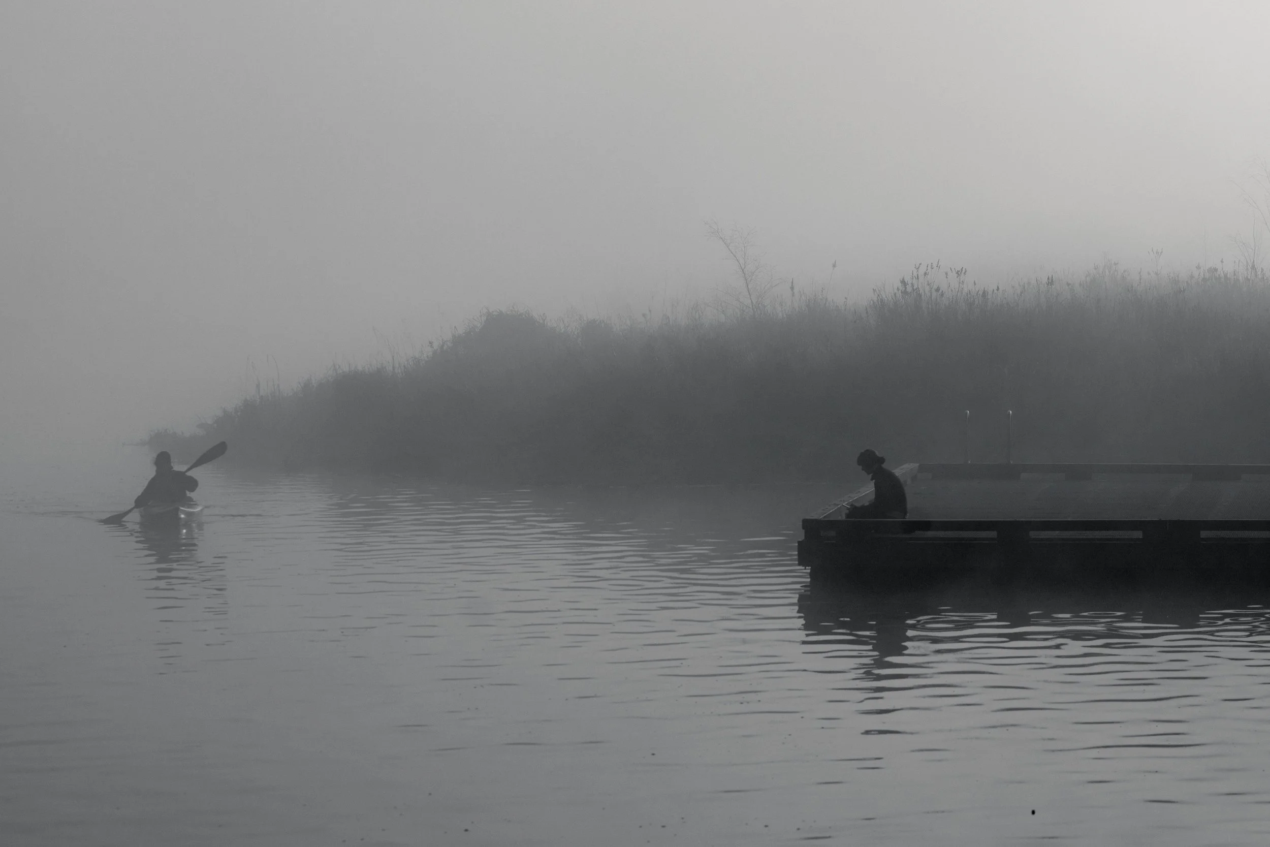 Two people are fishing on a foggy river, one sitting on a dock and the other paddling a kayak.