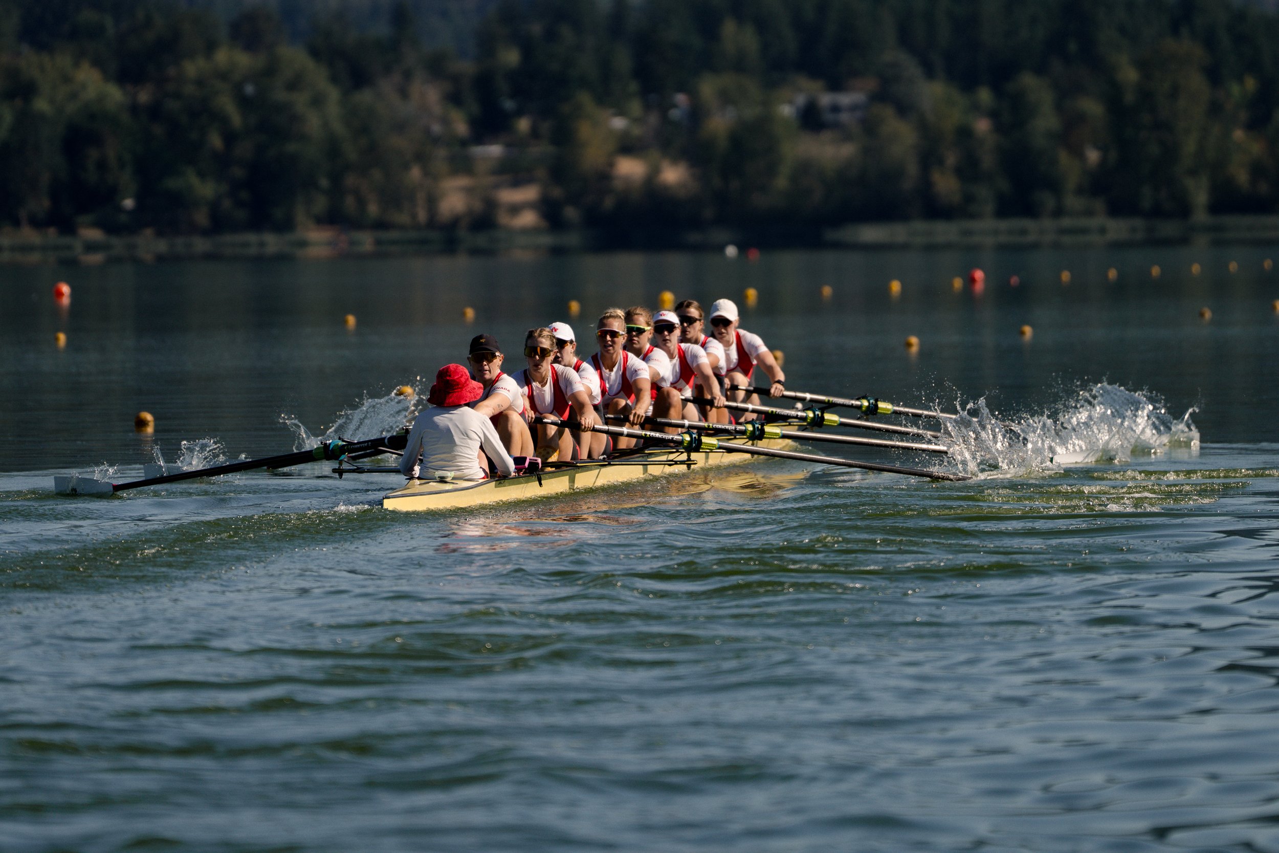 A team of eight women in matching athletic uniforms rowing a yellow boat on a calm body of water, with a person in a white shirt and red hat sitting at the back, surrounded by a natural landscape.