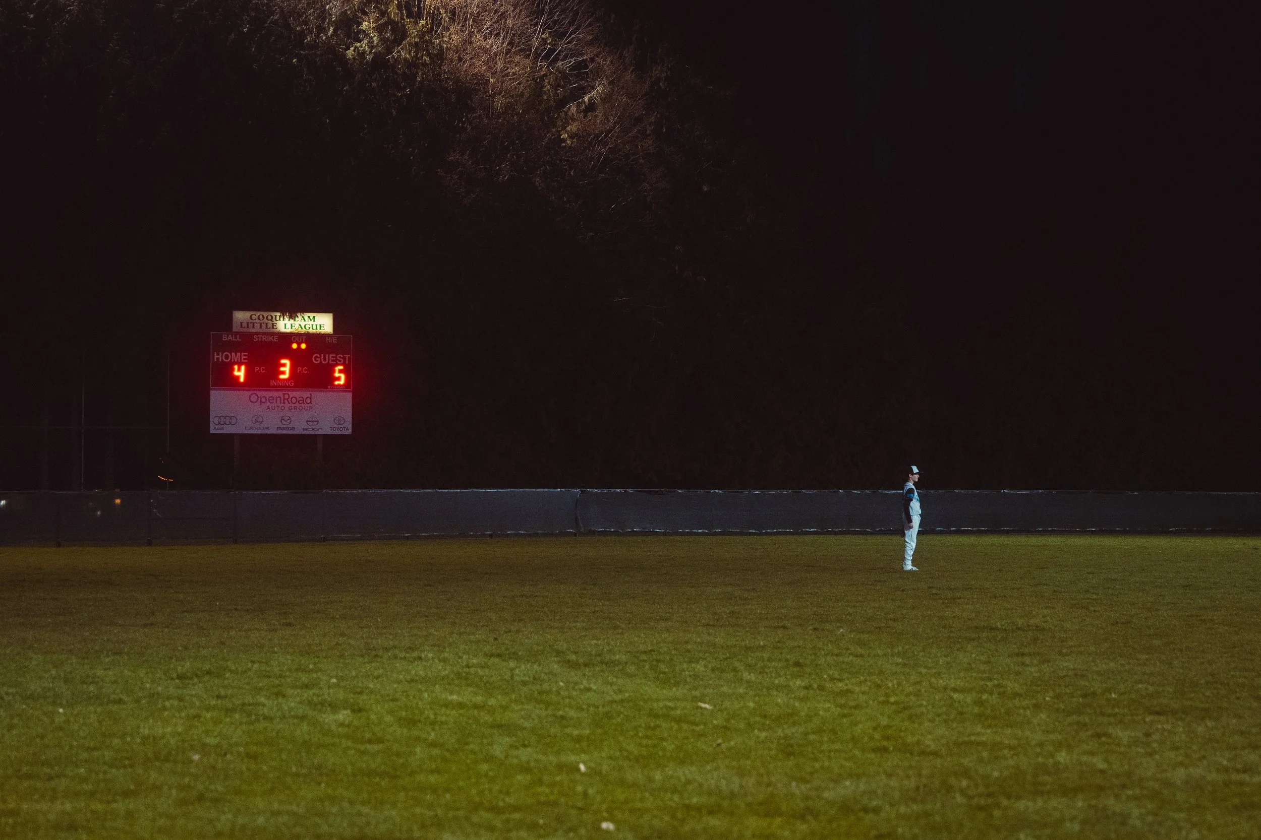 A nighttime scene at a sports field with a large illuminated scoreboard showing the score as Home 4, Guest 5, and the game in the third inning. A person in a cap and jacket stands on the grass field, and a row of trees is visible in the background.