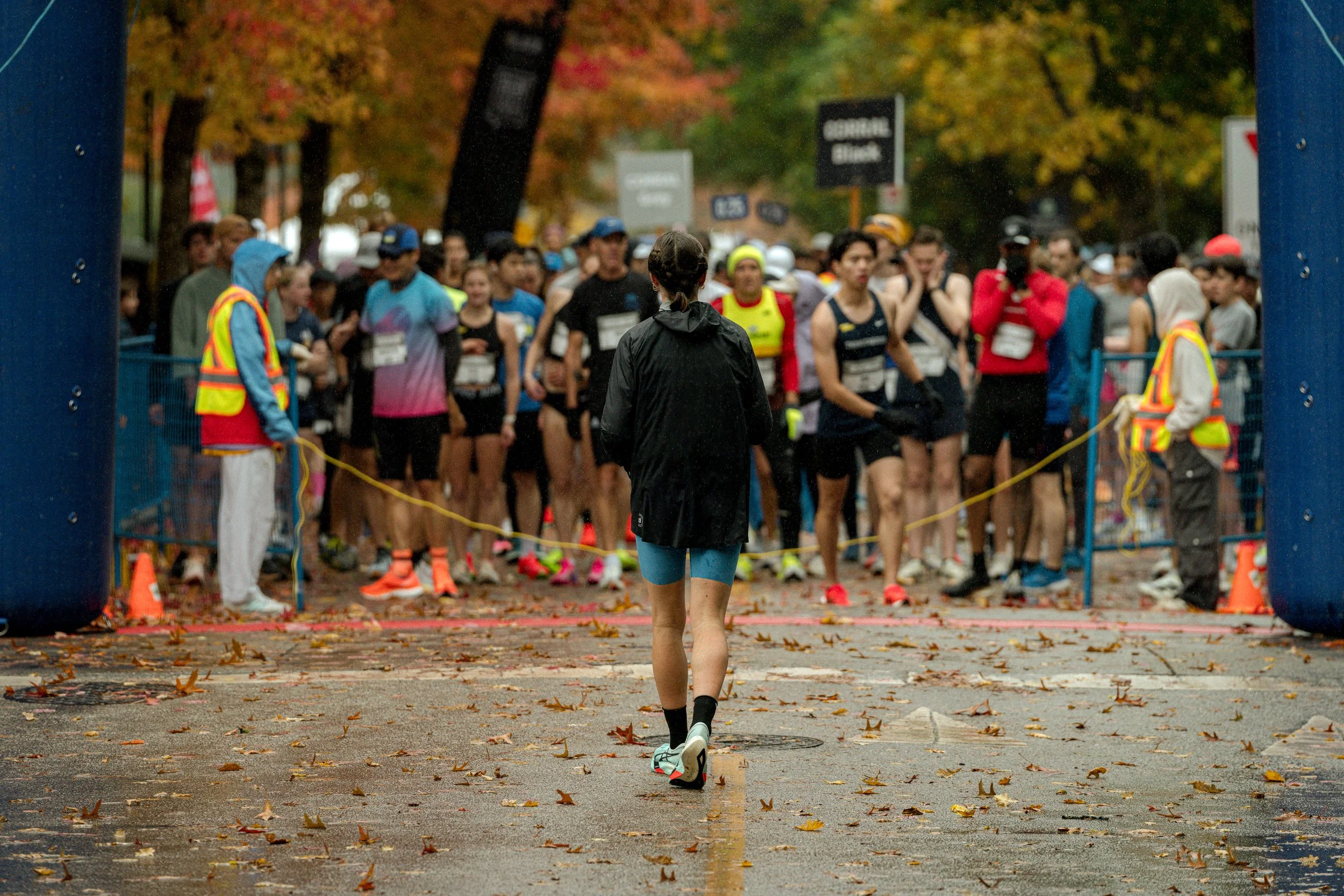 A group of runners gathered at the starting line of a race, with a woman standing in front facing them, in an outdoor setting with autumn-colored trees.